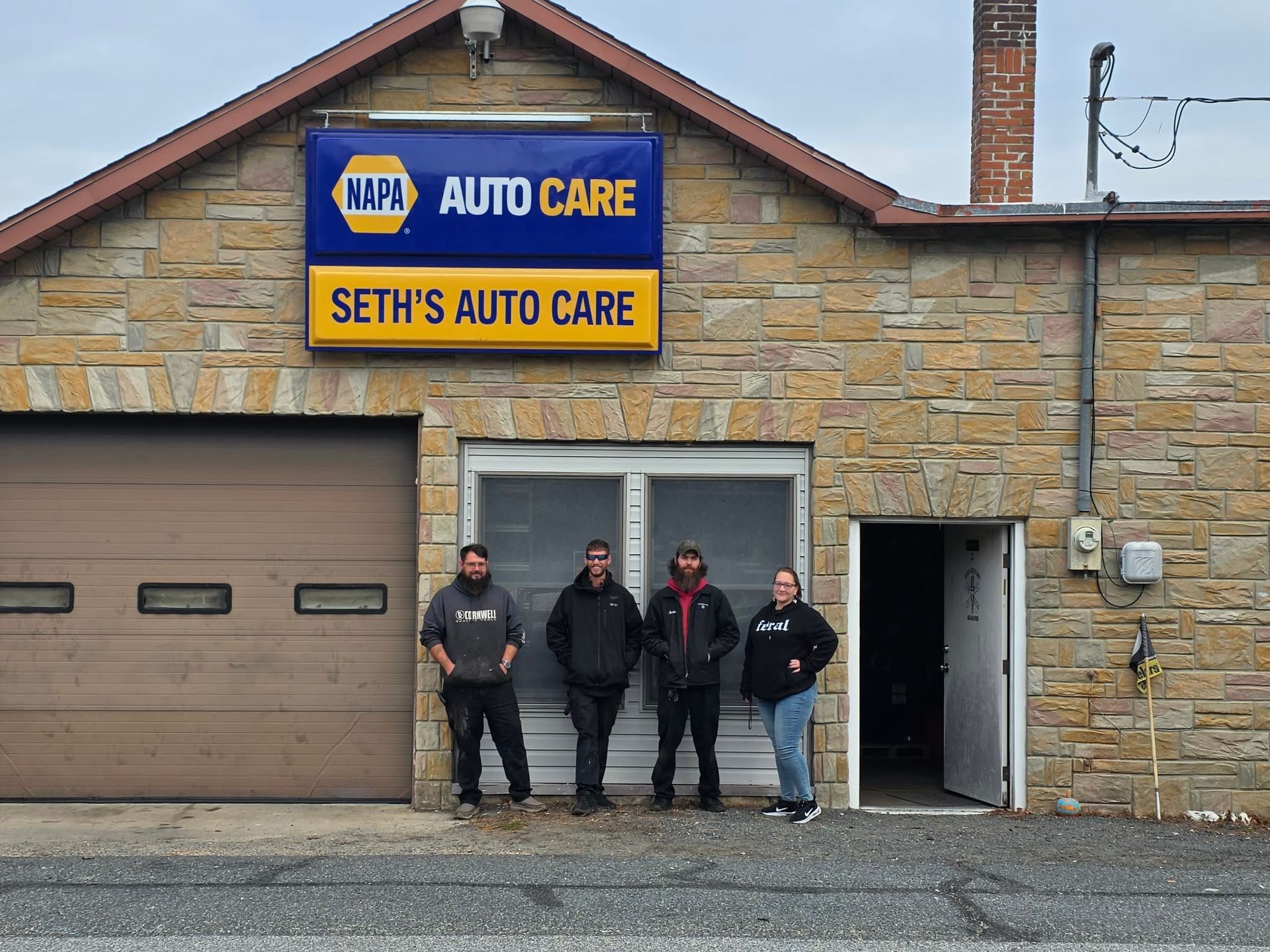 Four people stand in front of Seth's Auto Care, a stone building with a NAPA sign. Garage door and open side door visible. | Seth's Auto Care