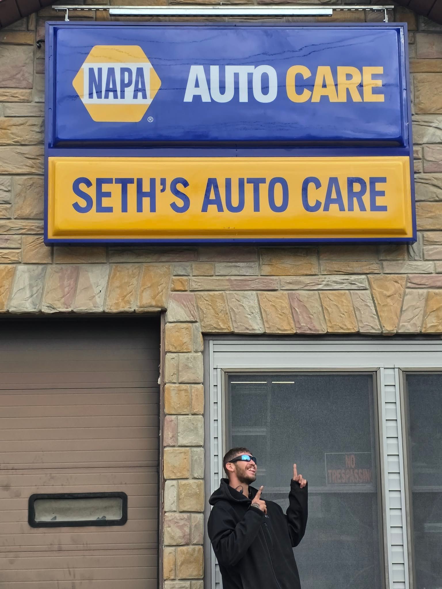 Sign for Seth's Auto Care, a NAPA Auto Care shop. Man points upward, wearing sunglasses, outside the shop. | Seth's Auto Care