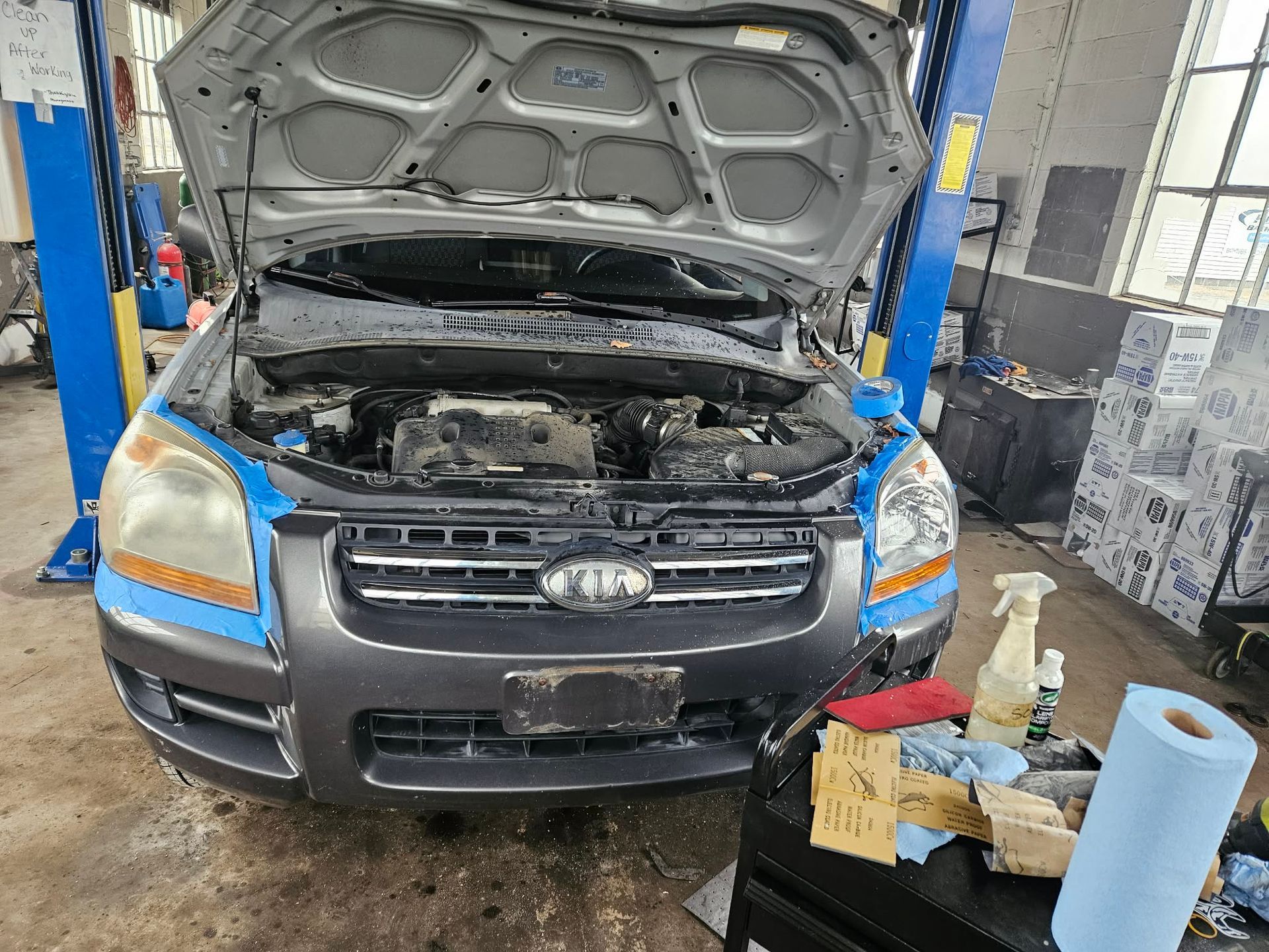 Car being repaired in a garage, hood open. Grey Kia Sportage, blue tape around the headlights. | Seth's Auto Care