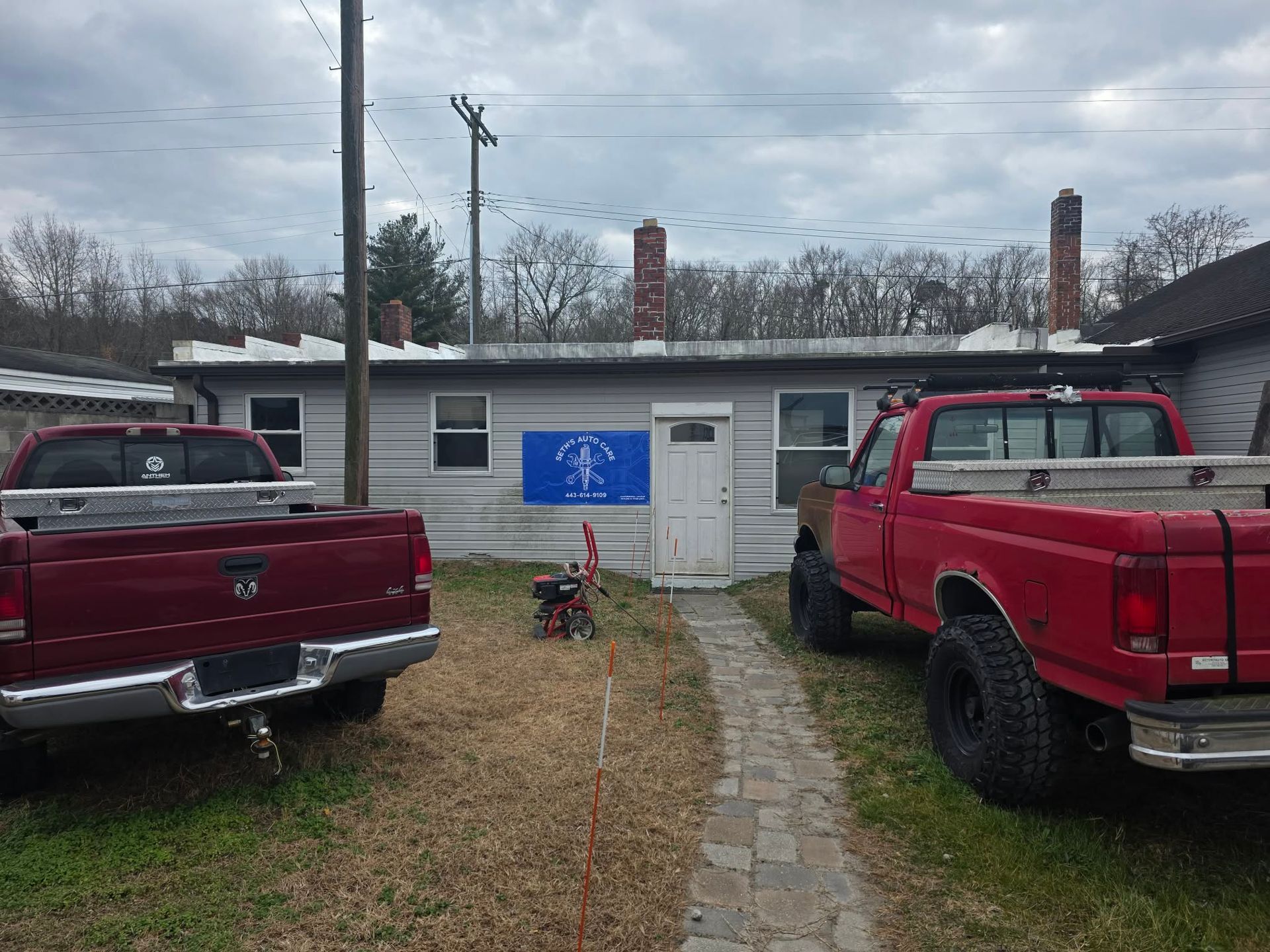 Two red pickup trucks flank a small building with a blue sign and a brick pathway. | Seth's Auto Care