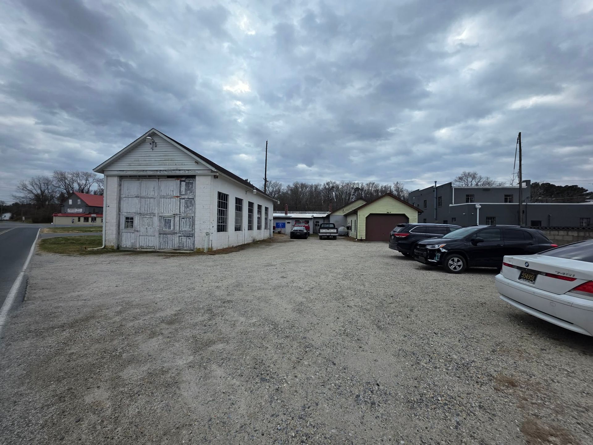 A gravel parking lot with several cars parked near a white building with large garage doors. Overcast sky. | Seth's Auto Care