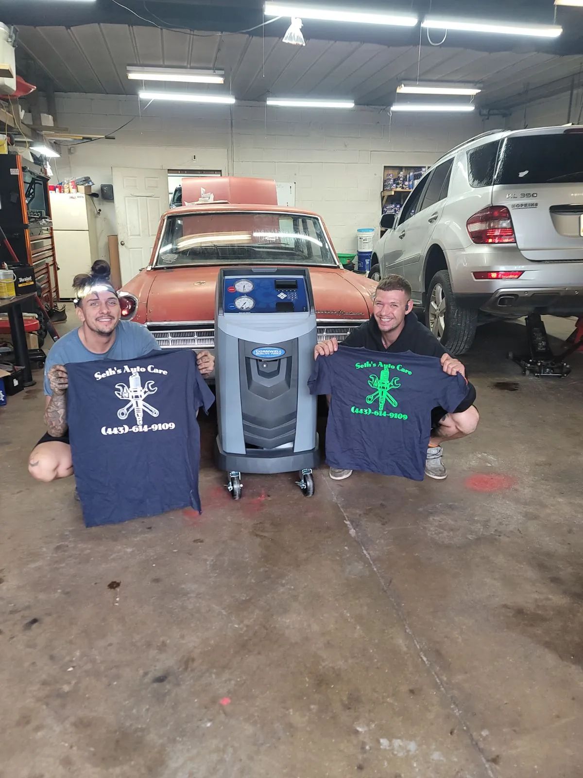 Two men in a garage holding shirts, flanking an AC recharge machine, with classic car and SUV in the background. | Seth's Auto Care