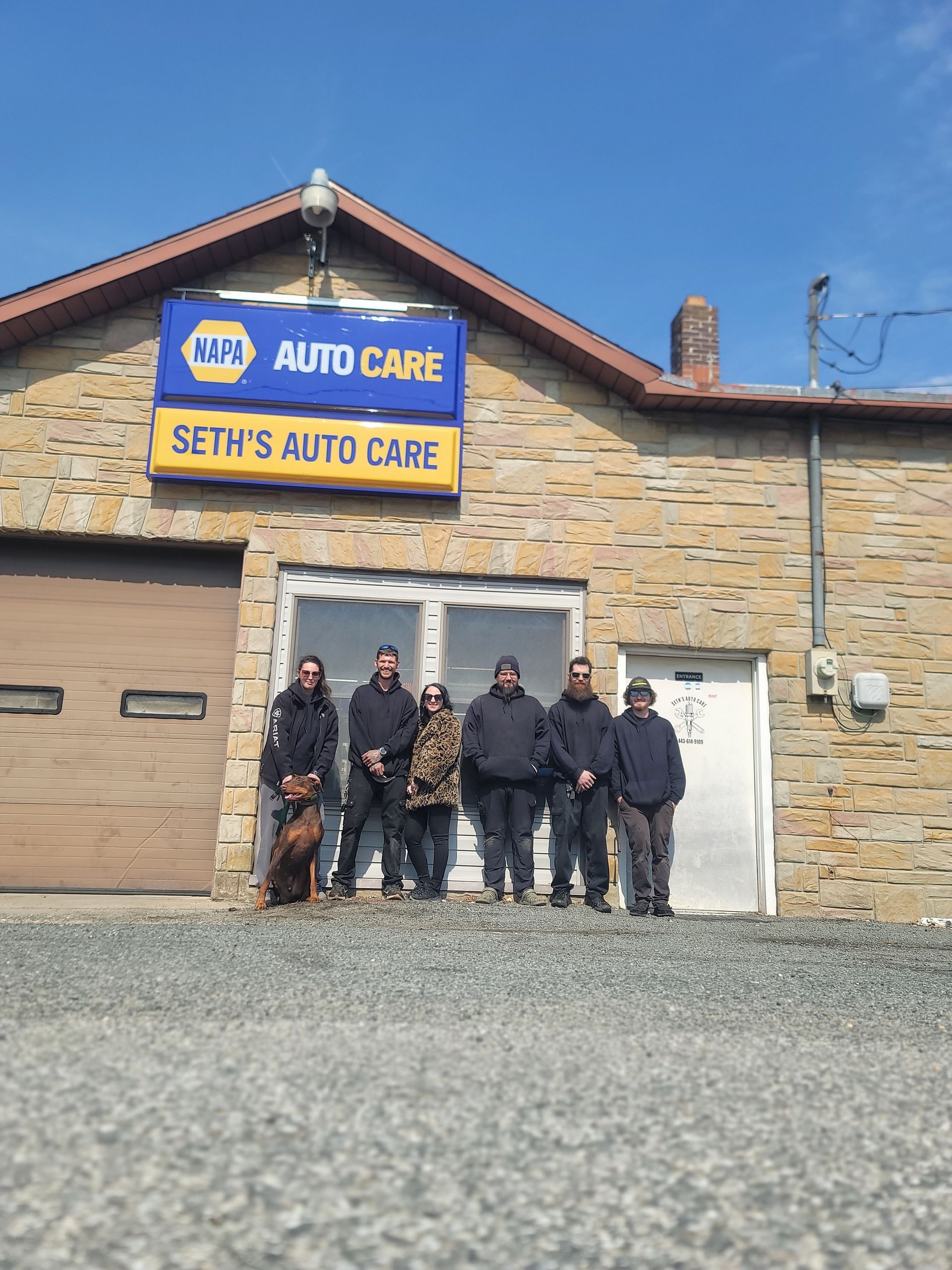 Two men in a garage holding shirts, flanking an AC recharge machine, with classic car and SUV in the background. | Seth's Auto Care