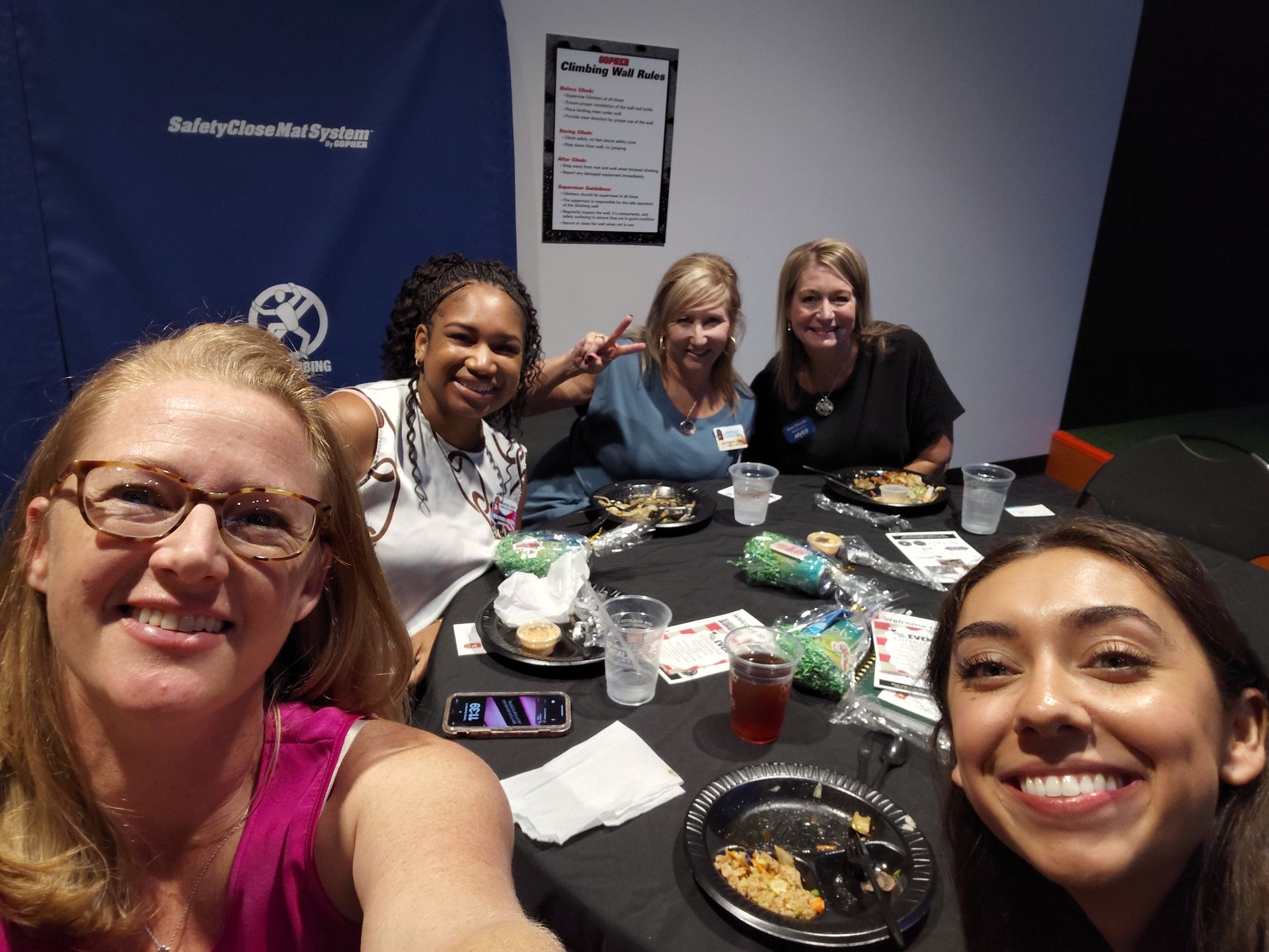 Five people smiling at a table with food, drinks. Room setting with a banner in the background.