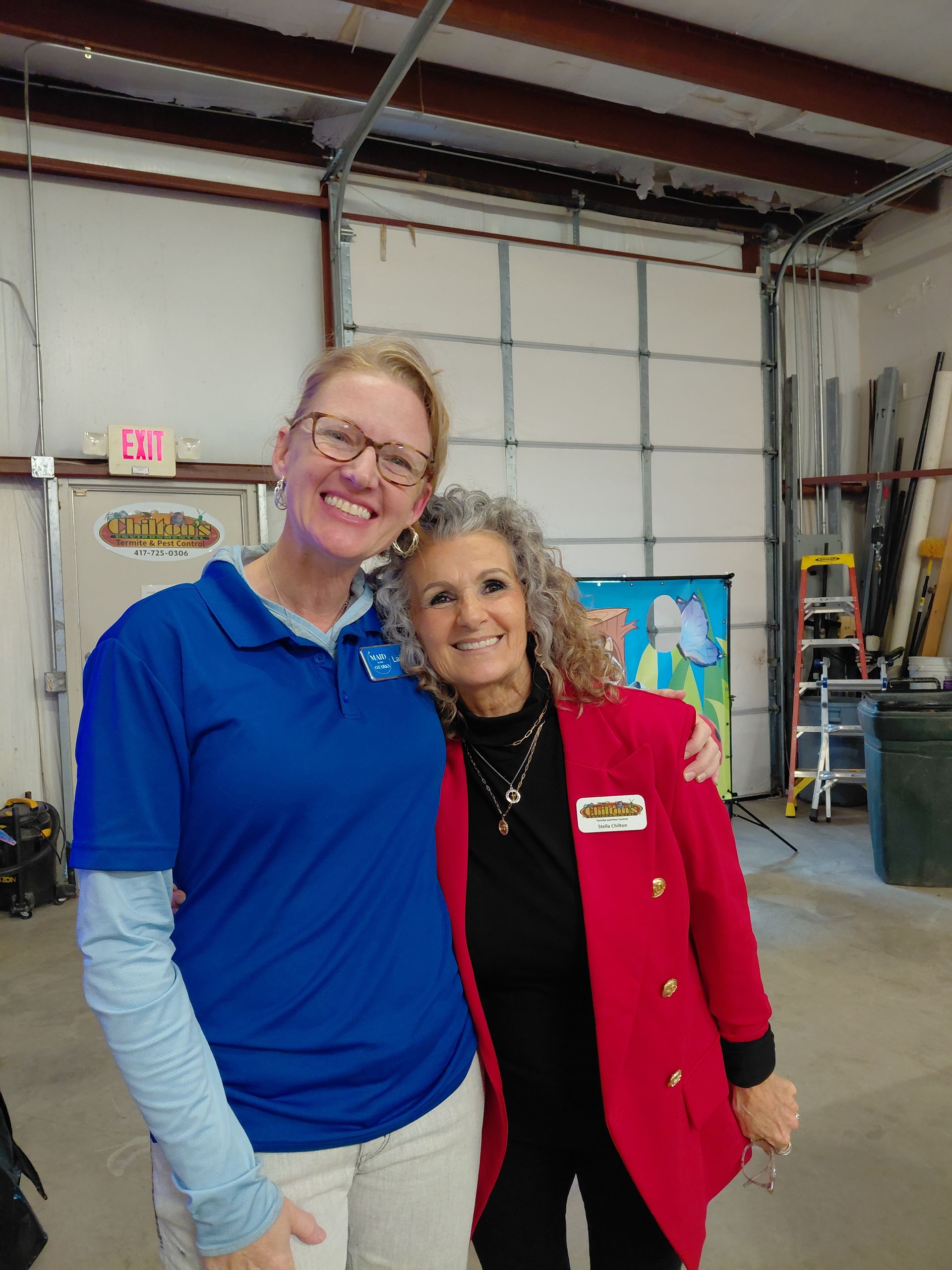 Two women smiling, arm in arm. One in a blue shirt, the other in a red blazer, in a workshop setting.