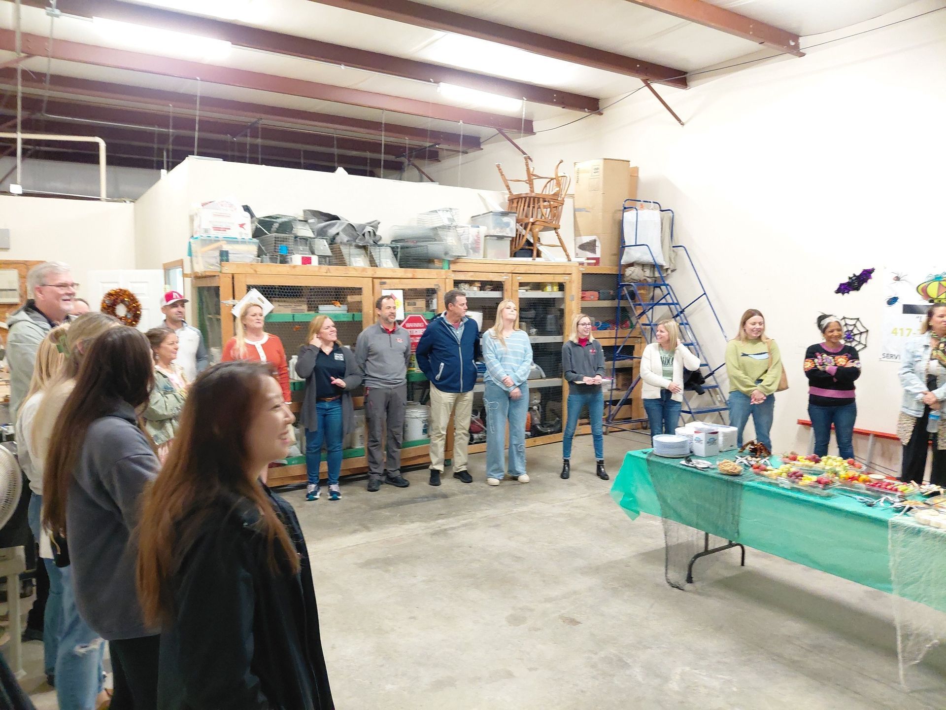 Group of people standing in a warehouse, some wearing party hats, gathered near a table.