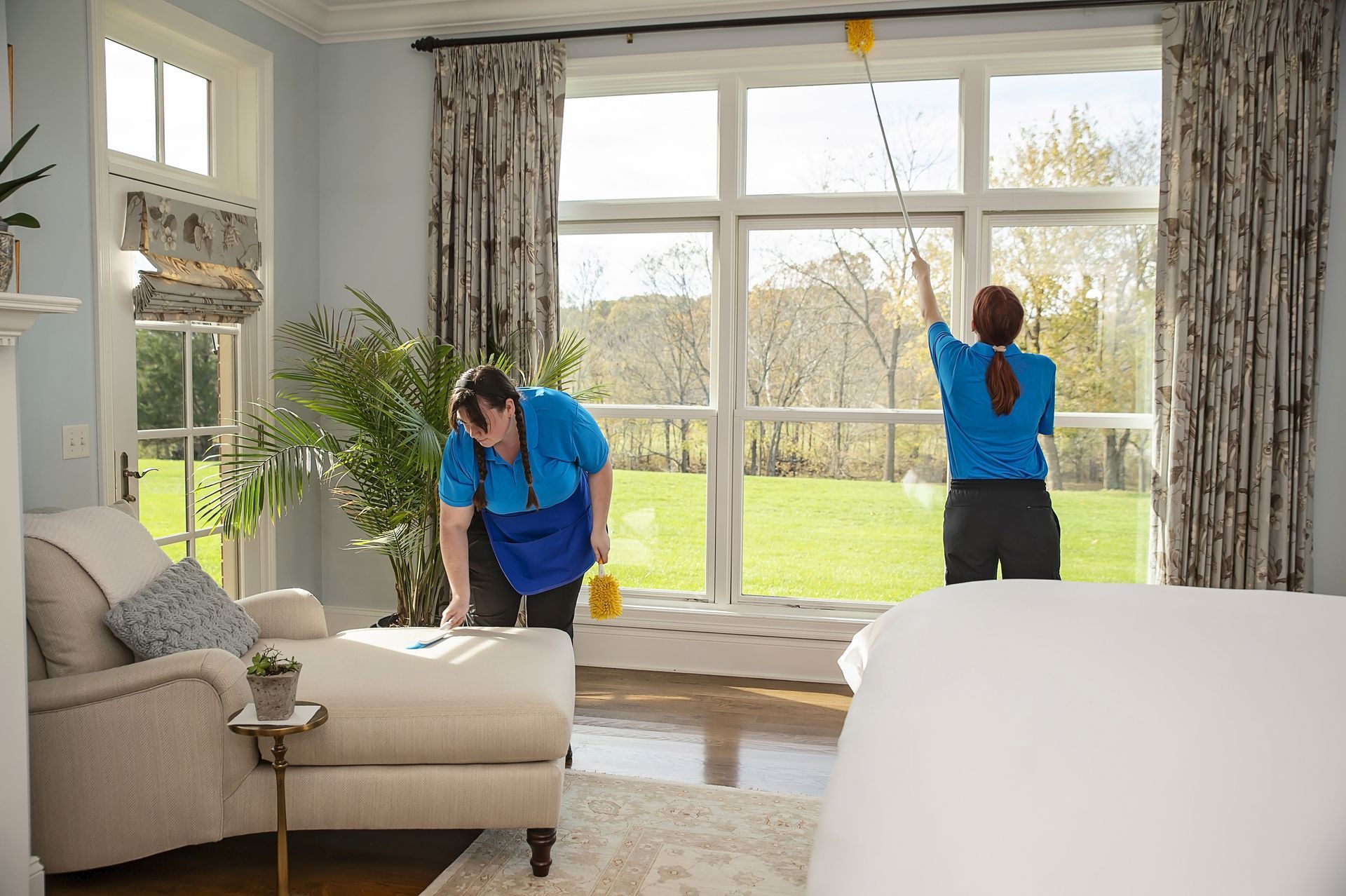 Two people cleaning a room with a large window. One wipes the window, the other cleans.