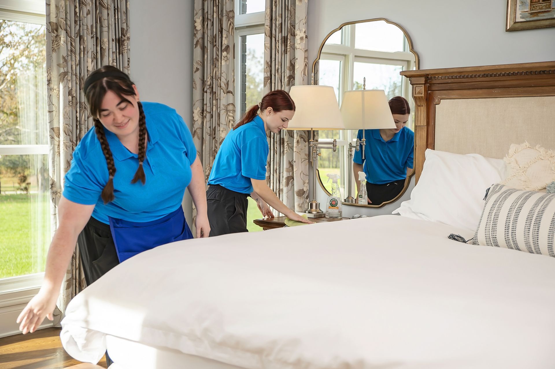 Two people in blue shirts making a bed in a well-lit bedroom.
