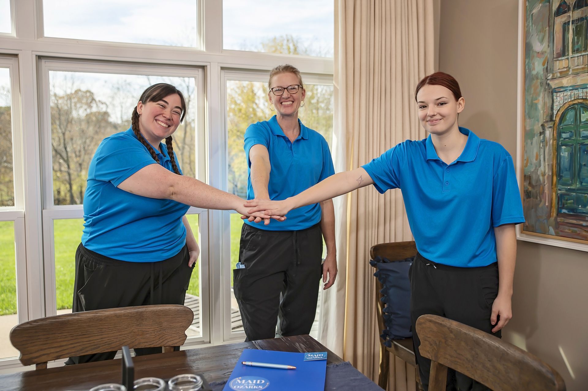 Three people in blue shirts, hands together, smiling. Standing by a window and table.