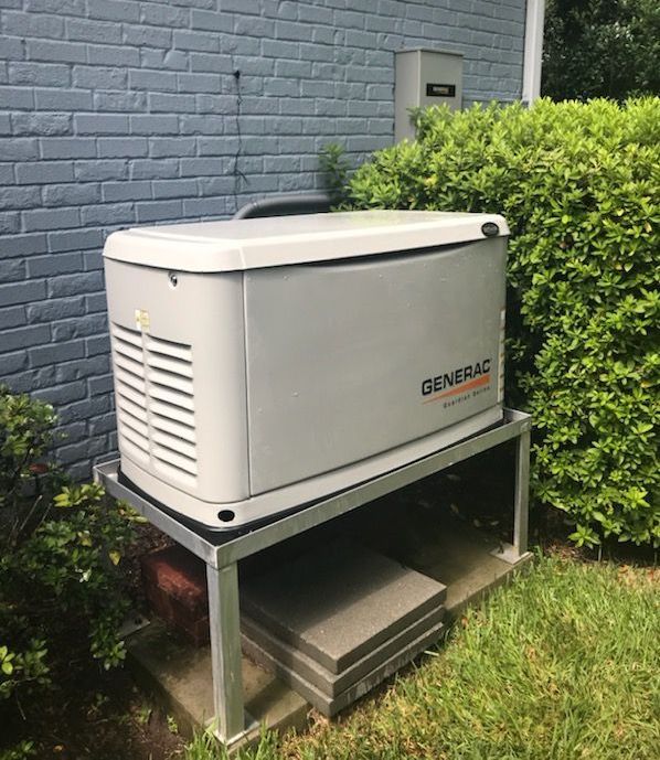 A Generac generator on a metal stand next to a brick wall and bushes.