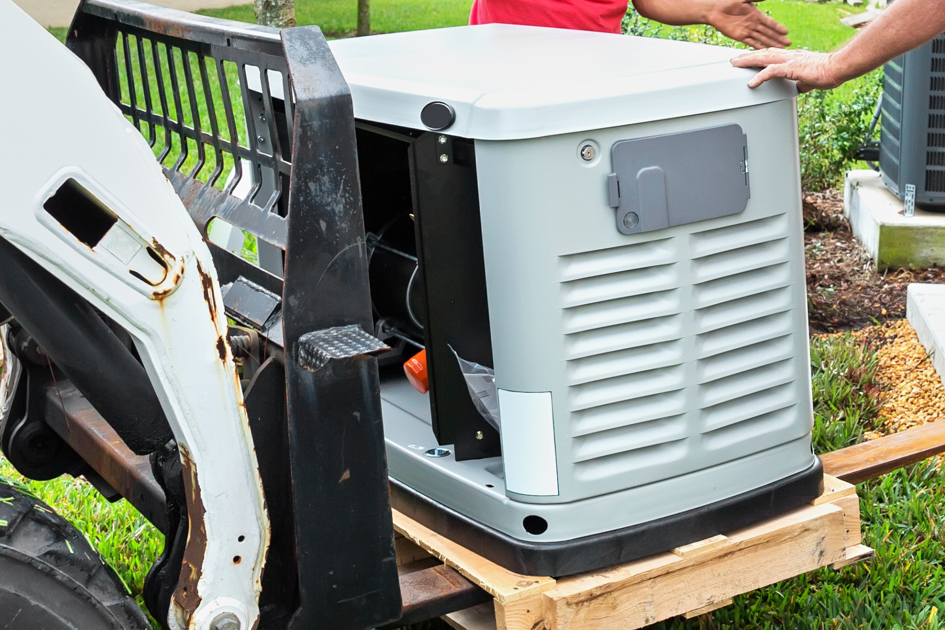 A generator on a pallet being moved by a small forklift. Two people's arms are visible. Outdoors, green grass.