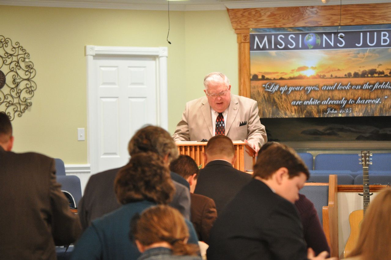 A man in a light suit preaches at a pulpit, addressing a seated congregation in a church during a 