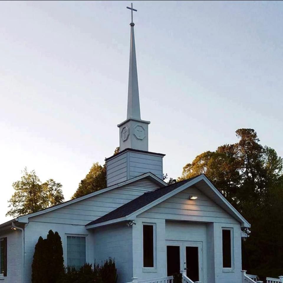 White church building with a tall steeple and cross against a pale blue sky.
