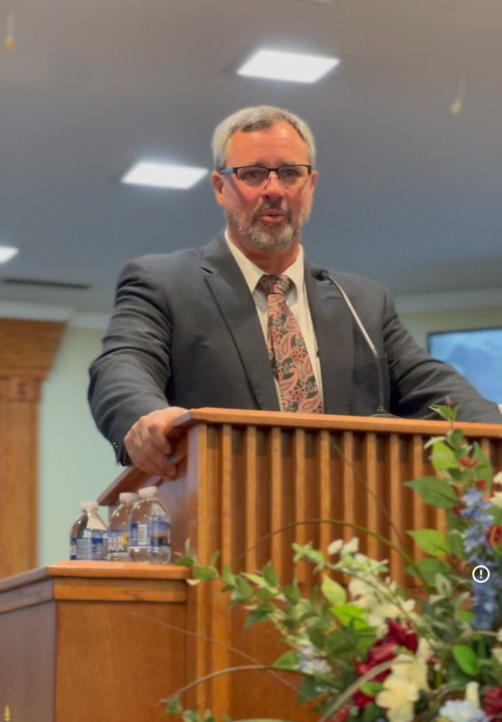 Man with glasses and a gray beard giving a speech at a podium, wearing a suit and tie. Flowers are in the foreground.