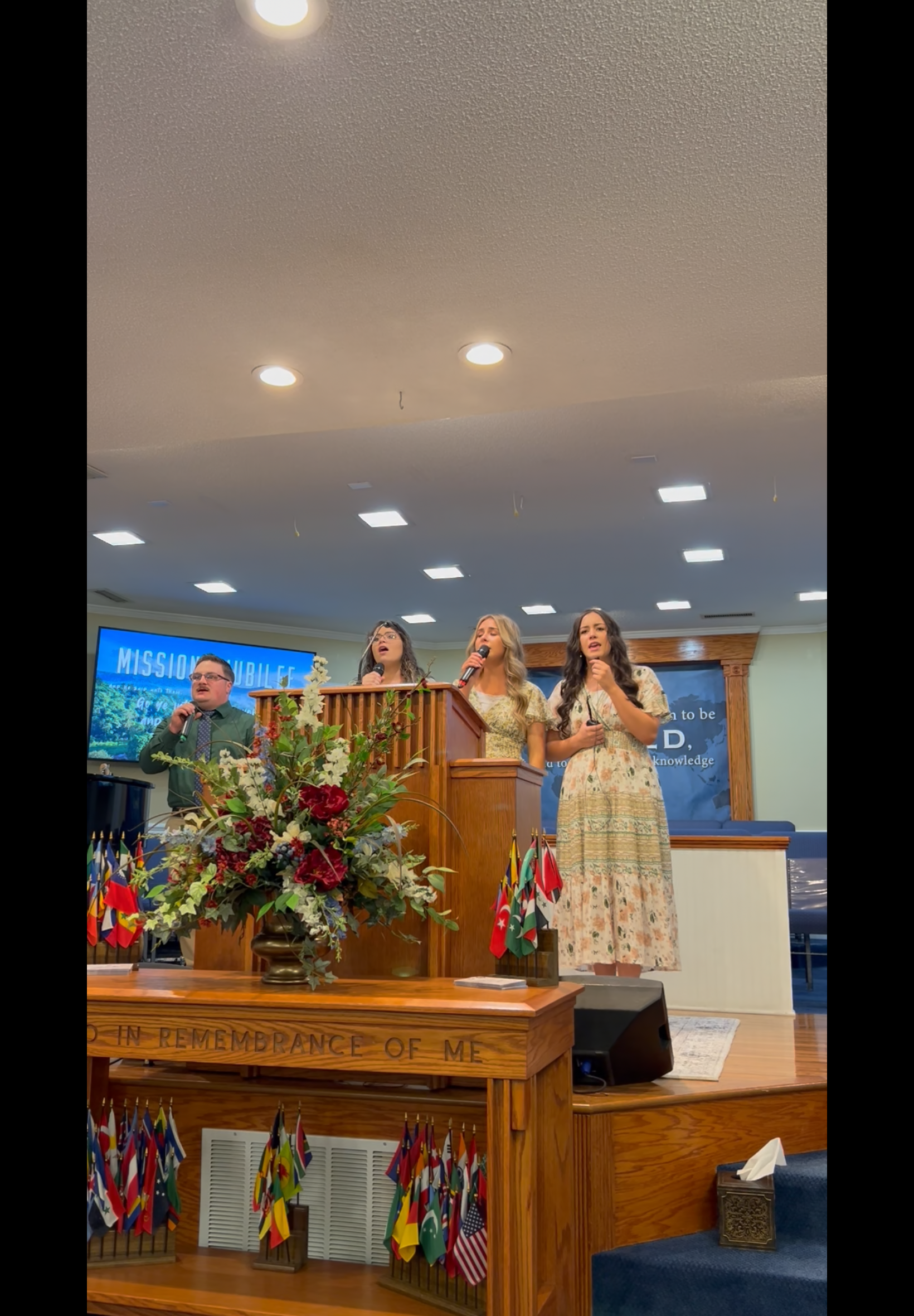 Three women singing in a church service, standing on a stage. The women wear dresses, with one using a microphone.