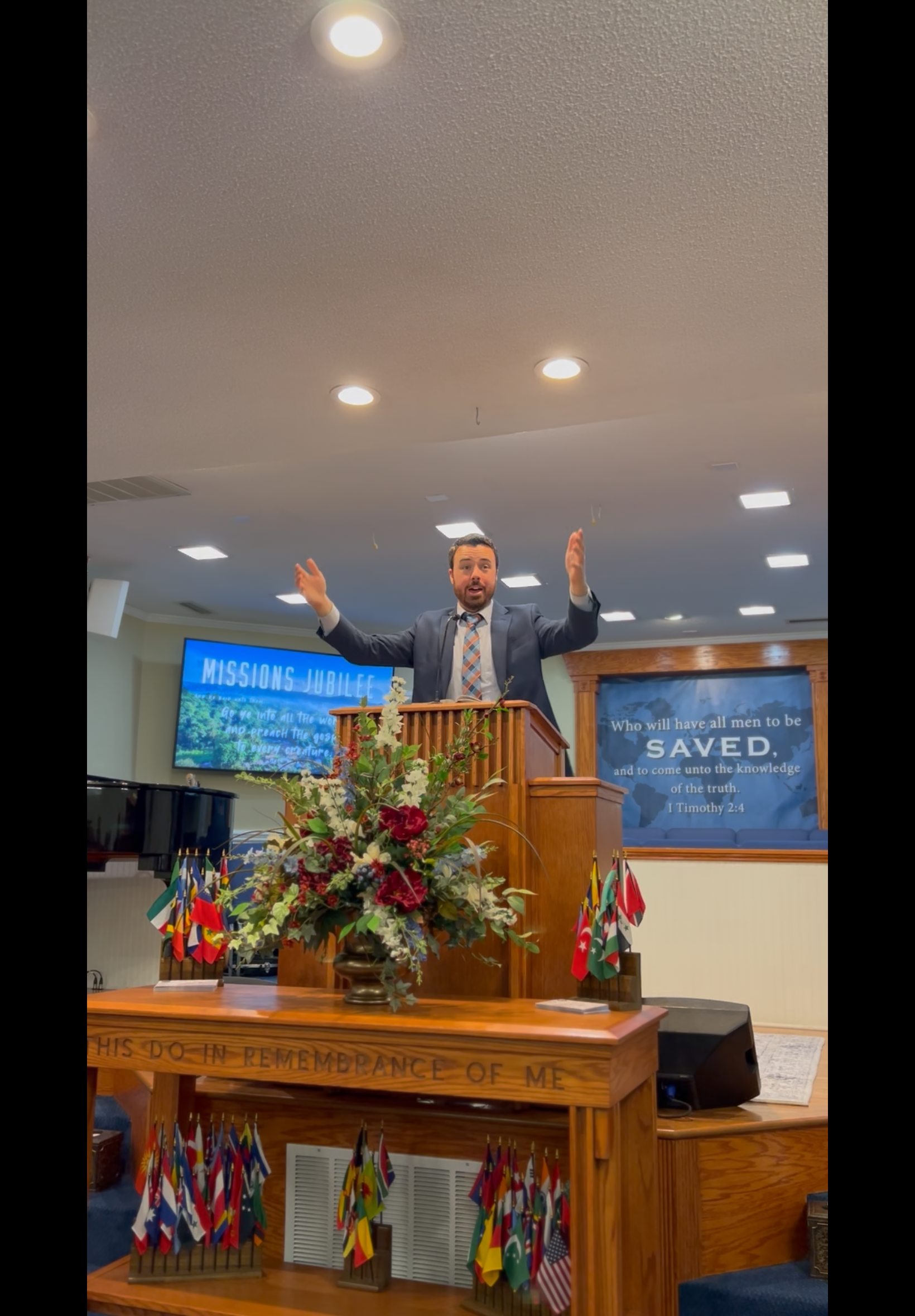 A man in a suit with arms raised preaching from a wooden podium in a church. Flags and floral arrangements are on the podium.