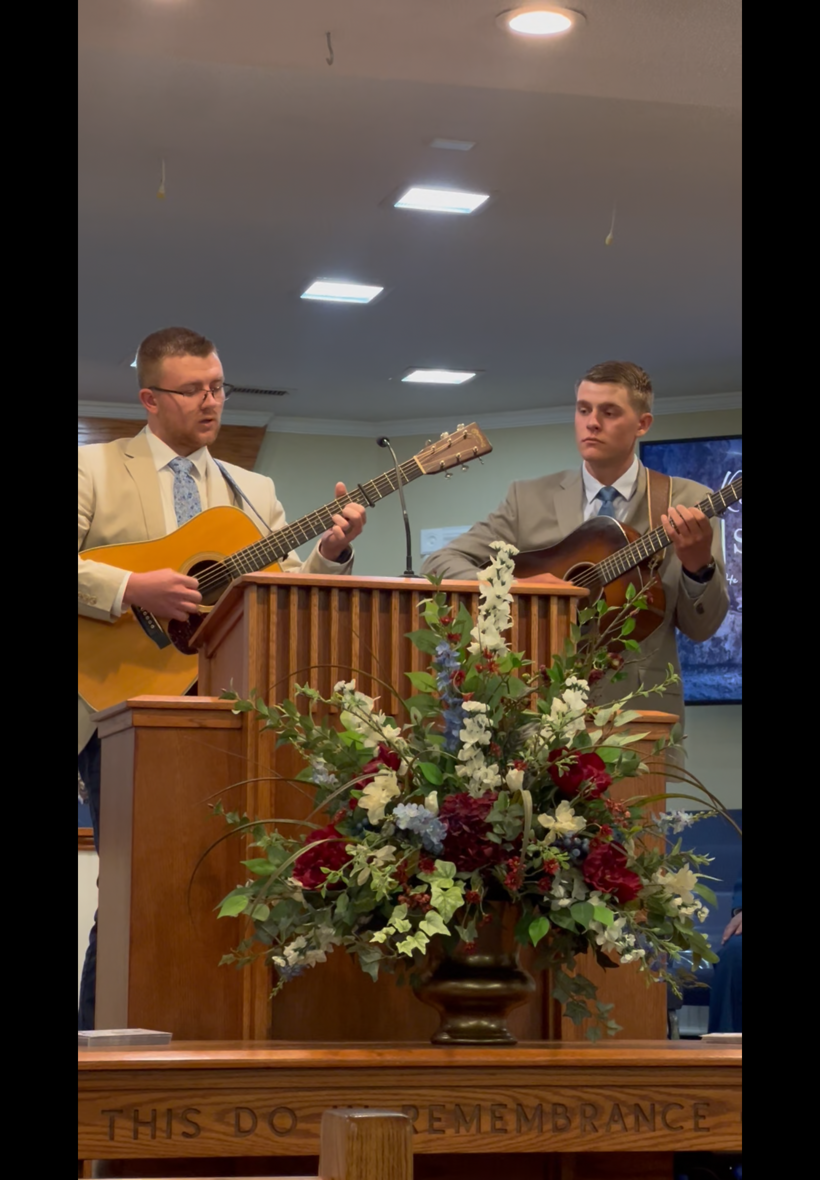 Two men in suits play acoustic guitar and mandolin at a church pulpit with flowers. They are singing.