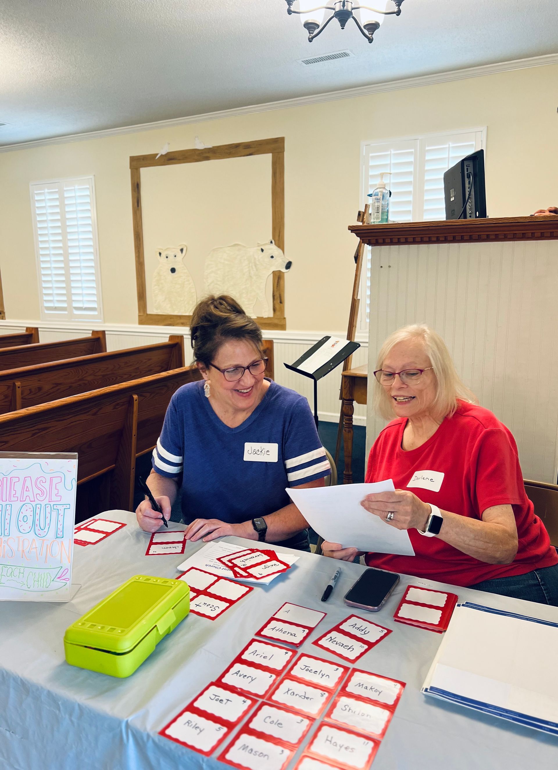 Two women smile while organizing name tags at a table inside. The woman on the left writes, the other holds a paper.