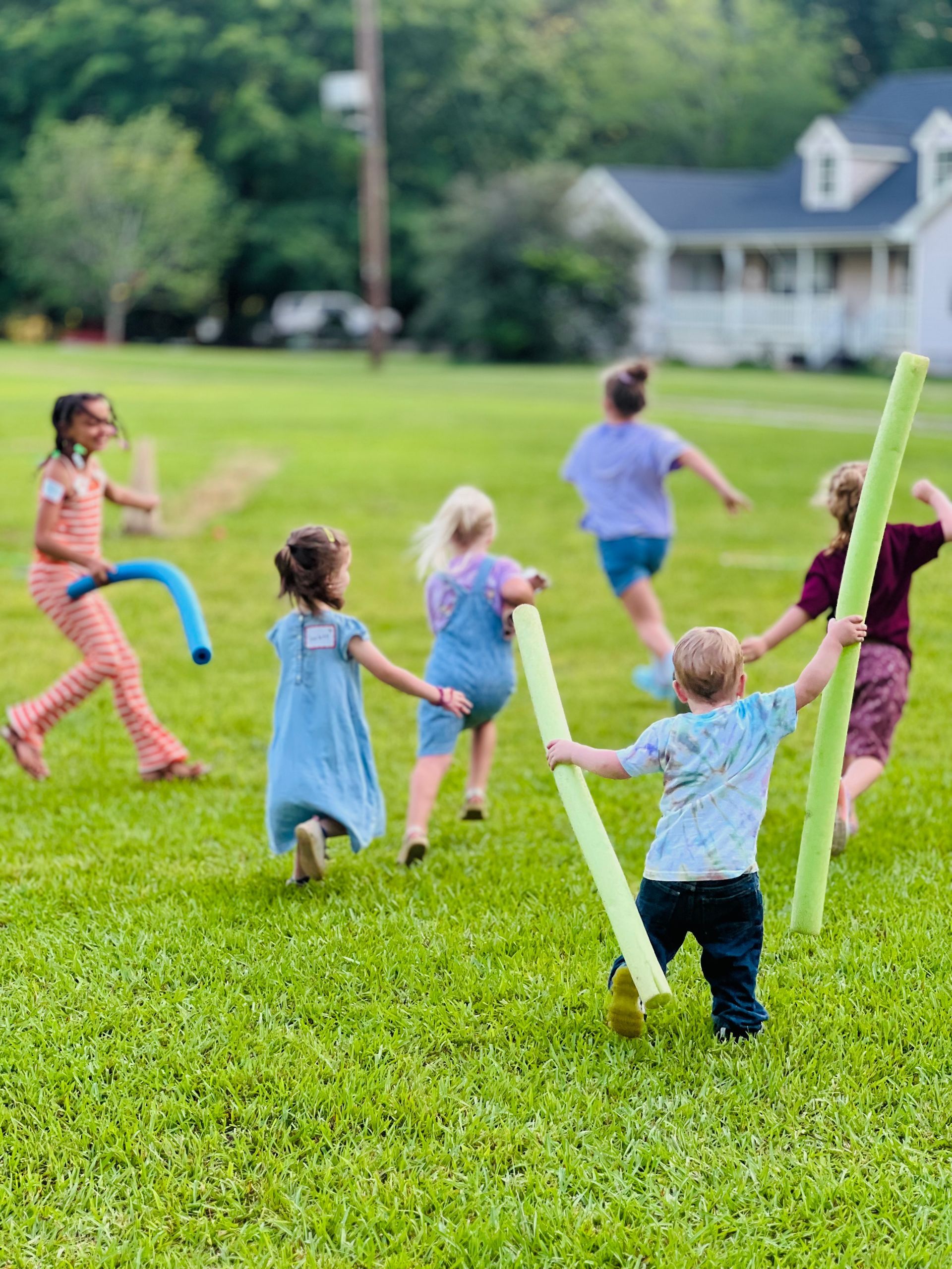 Children playing a game in a grassy yard, using pool noodles.