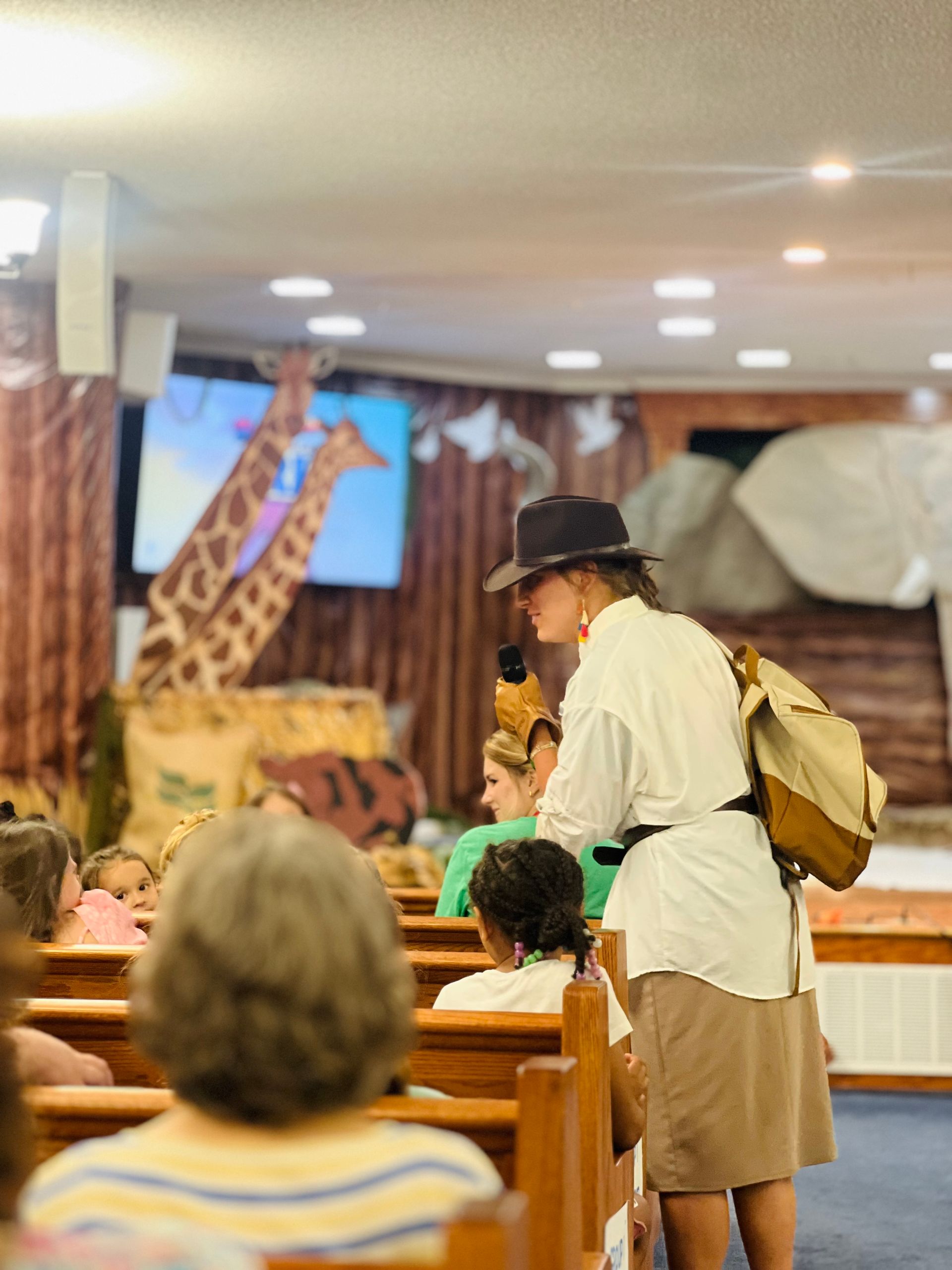 Person in safari attire stands in a church, possibly leading a children's event such as Vacation Bible School.