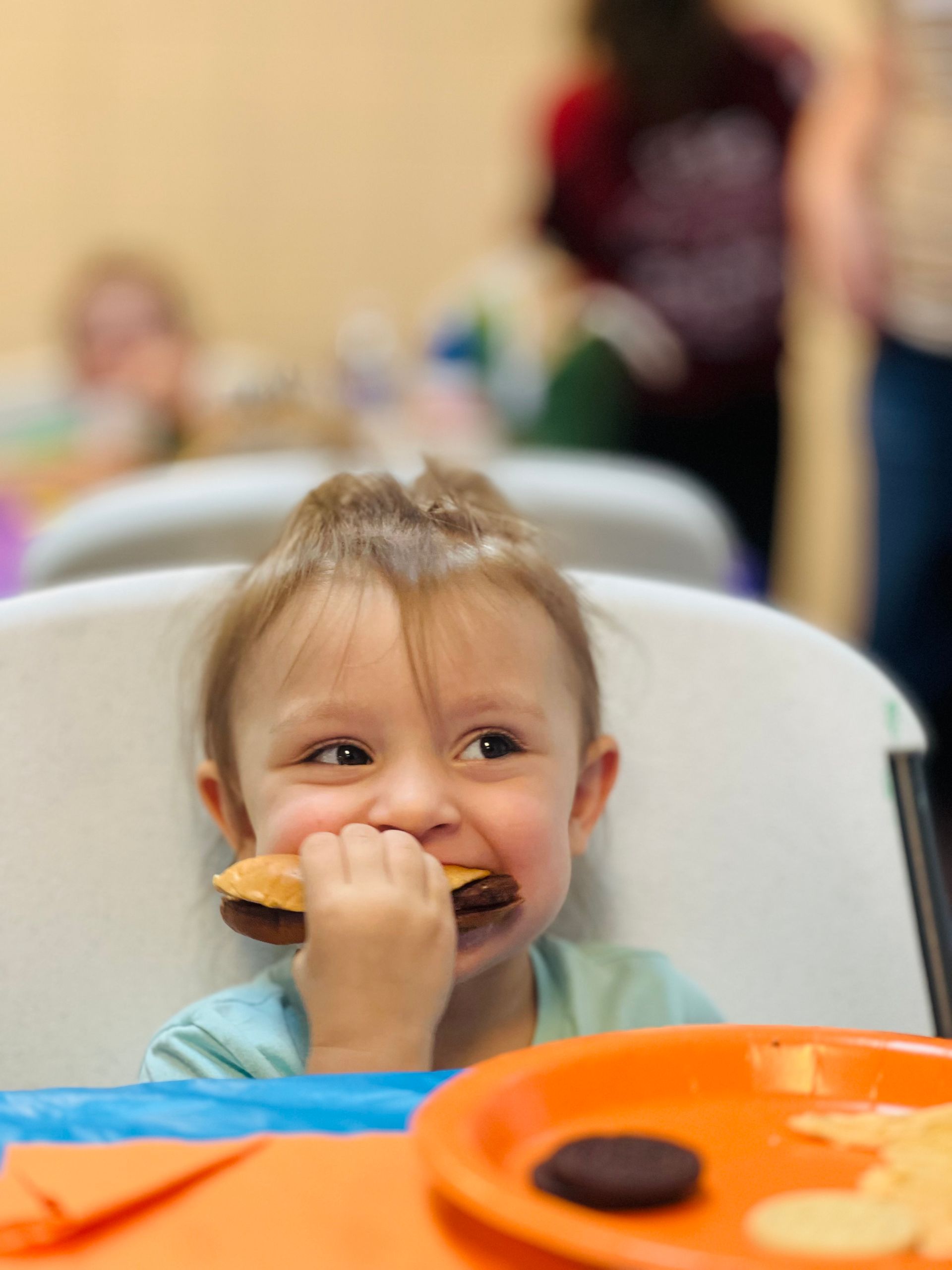 A young child happily bites into a chocolate cookie, sitting at a table. The child's expression is joyful.