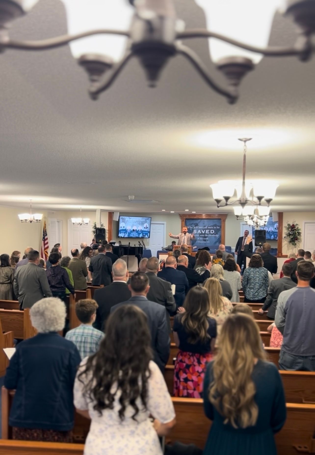 People standing in a church, facing a stage with a preacher. Neutral colors, chandeliers hang from the ceiling.