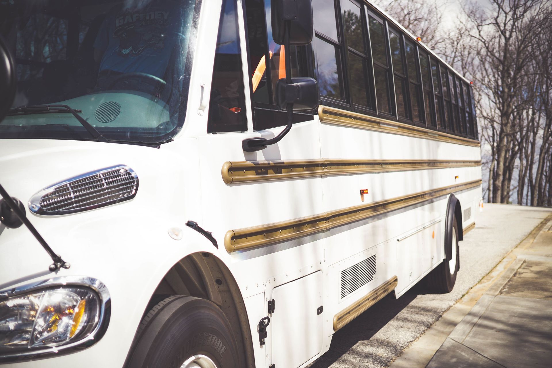 White school bus parked on the side of a road with bare trees in the background.