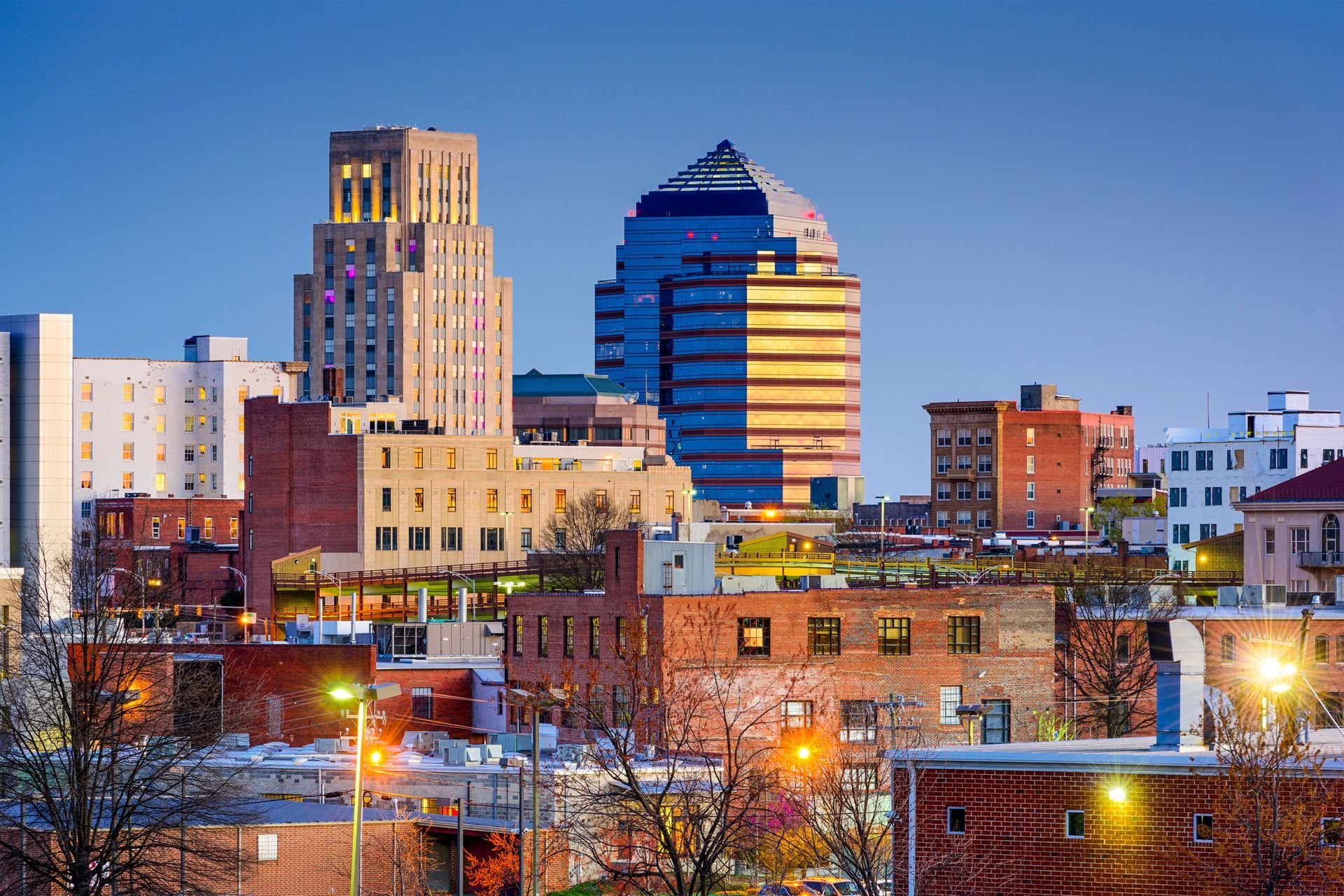 City skyline at dusk with brick buildings, lit windows, and modern skyscrapers against a clear blue sky.