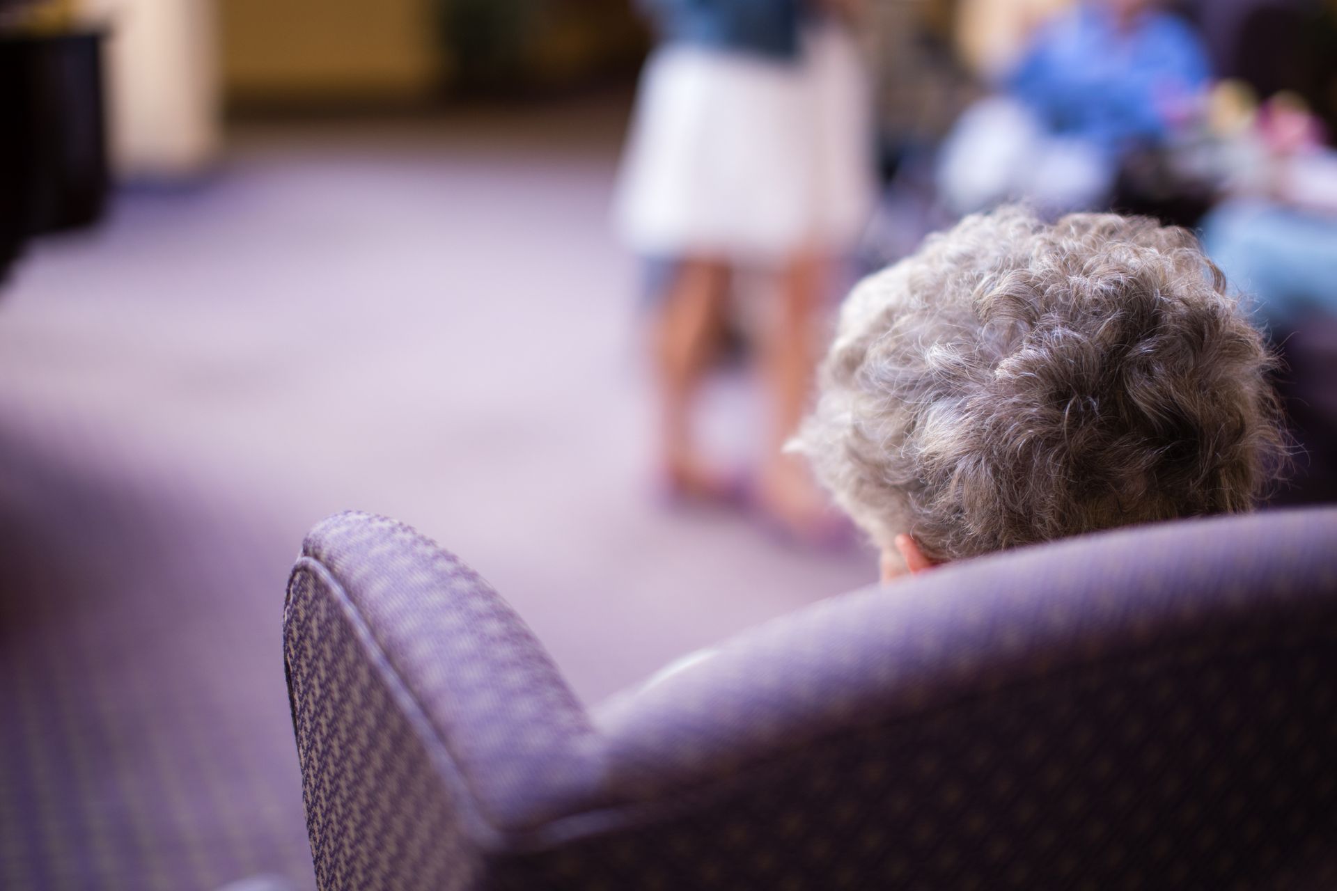An elderly person with gray hair sits in a purple armchair, watching a group of people.
