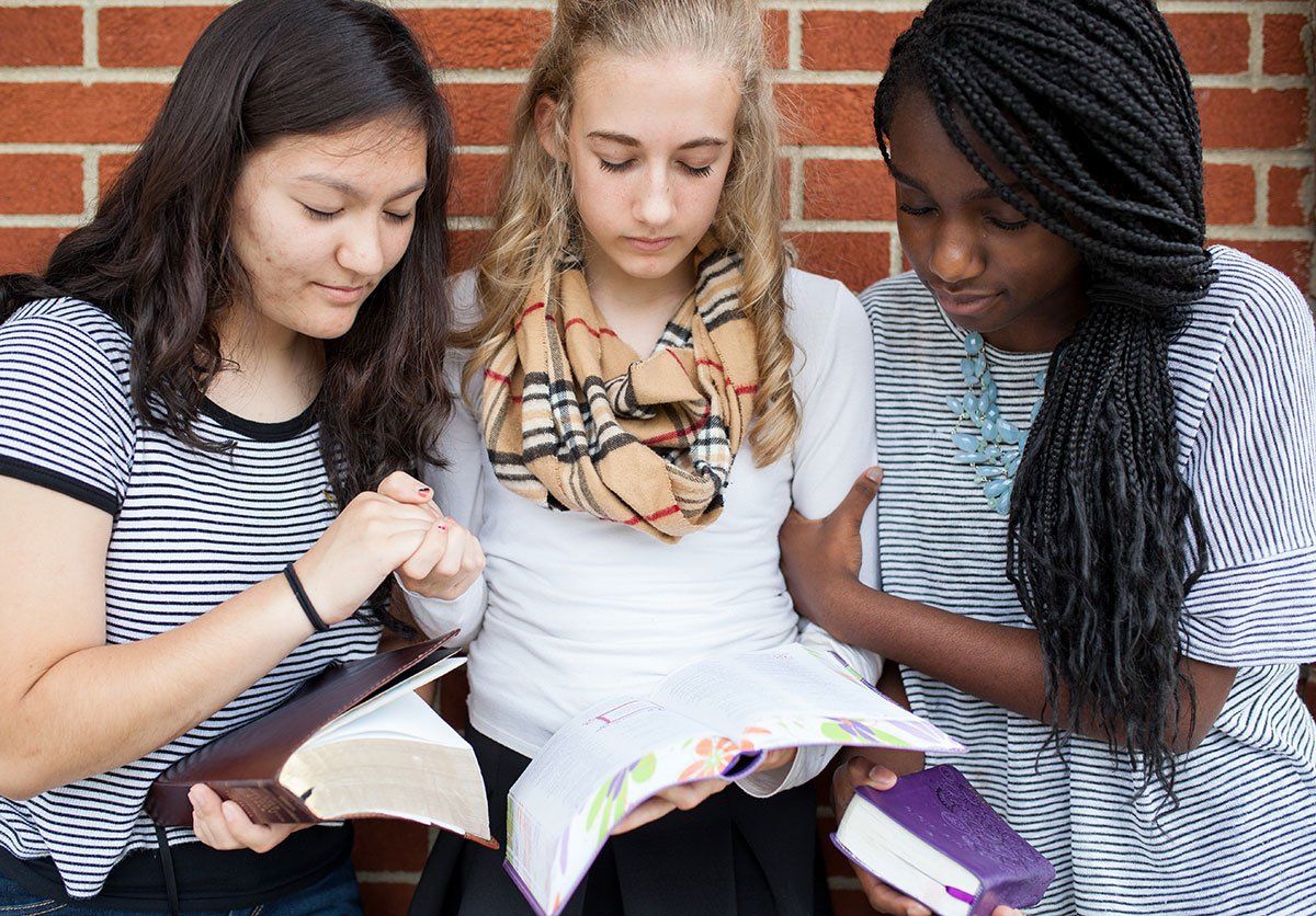 Three young women reading Bibles and praying together in front of a brick wall.