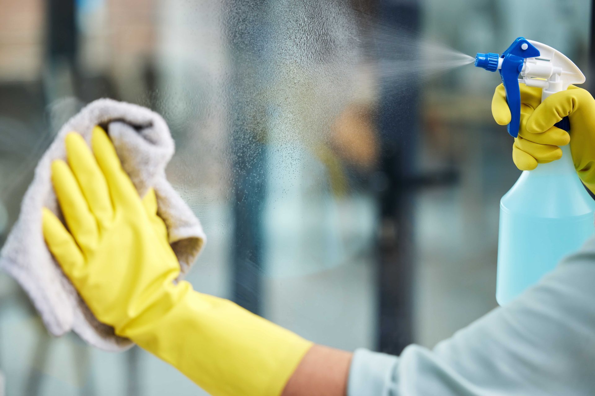 A person wearing yellow gloves is cleaning a window with a cloth and spray bottle.