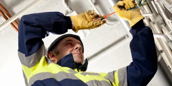 Electrician in yellow gloves and reflective jacket working on overhead pipes.