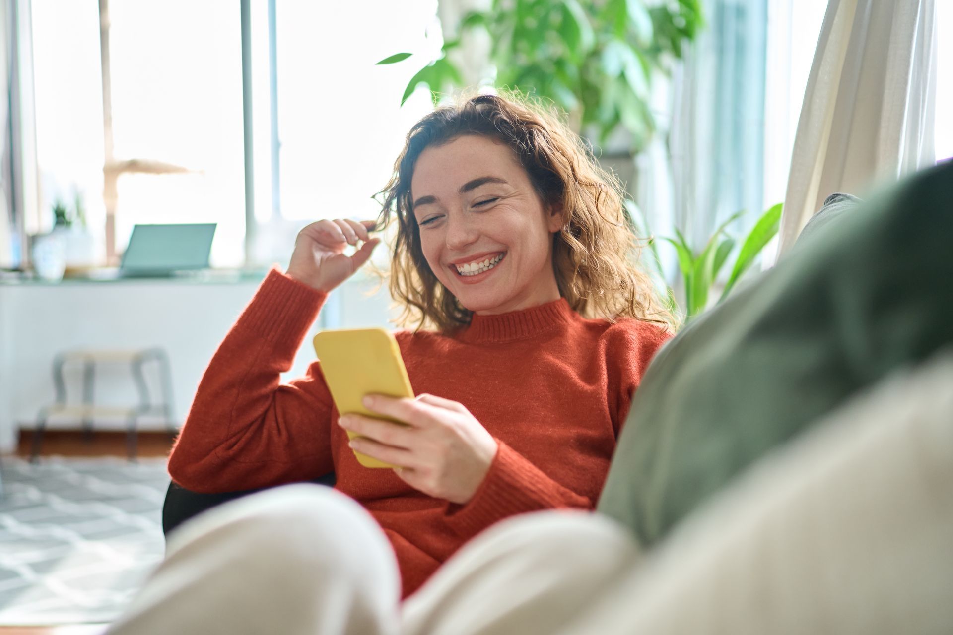 A person in a rust-colored sweater smiling while looking at a yellow smartphone, sitting on a couch in a bright room.