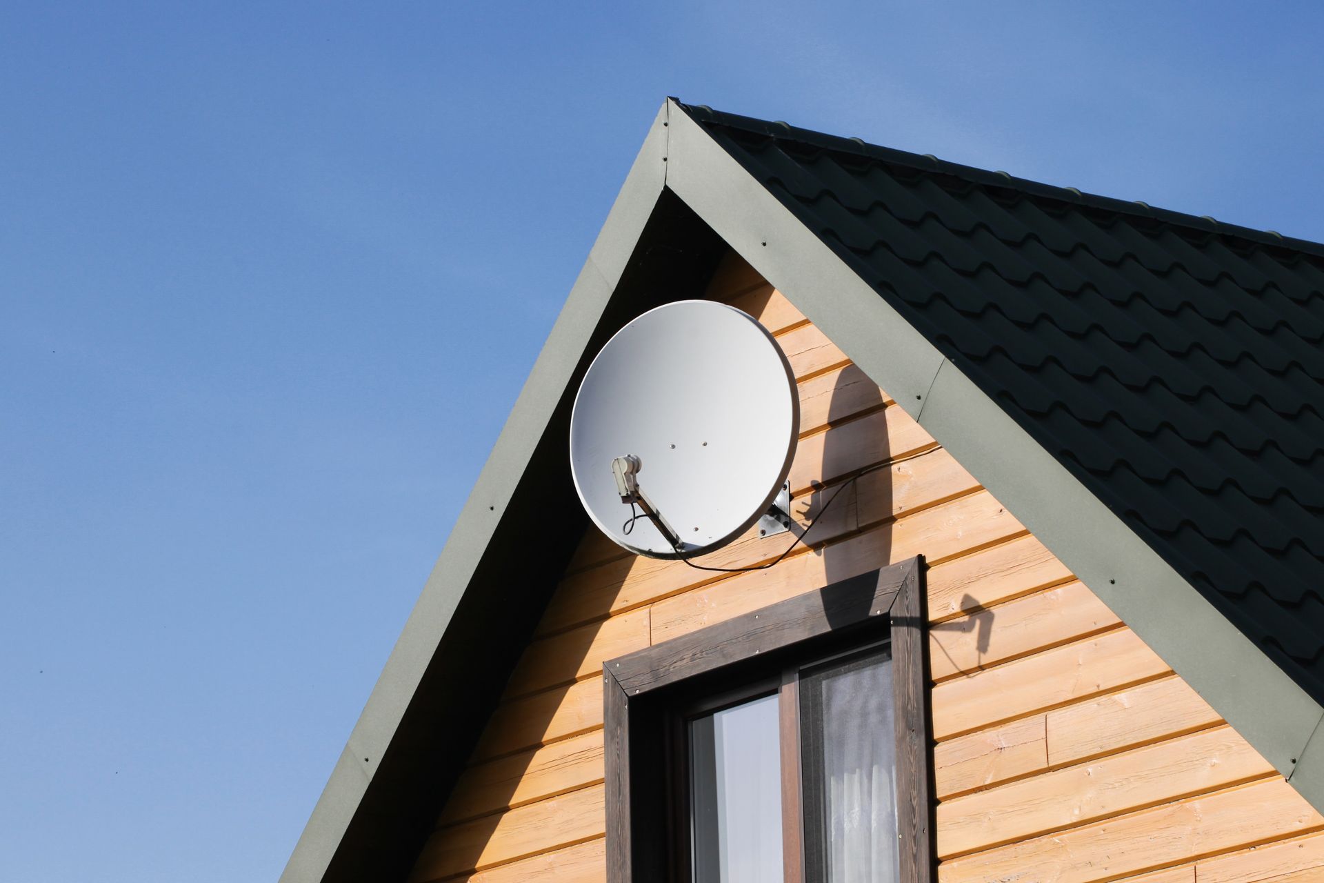 A satellite dish is mounted on the light-colored wooden gable wall of a house with a dark shingled roof against a blue sky.