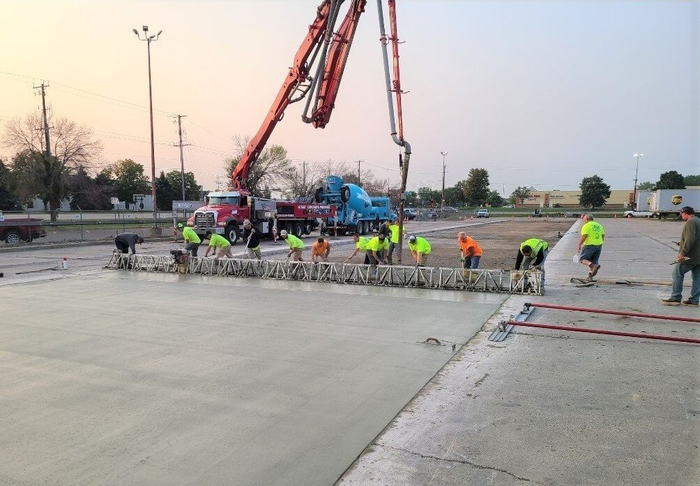 Team of men wearing neon shirts working together to level out a large mass of concrete with concrete delivery and pouring machines in the background.