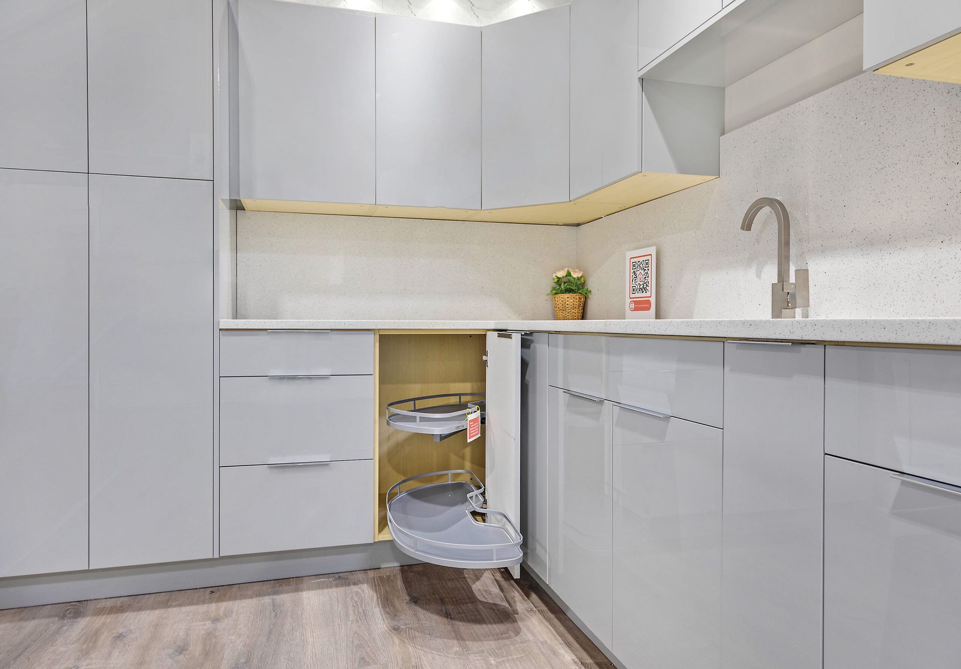 Light gray kitchen cabinets with a pull-out trash can. Stainless steel faucet, light countertop, and wood floor.