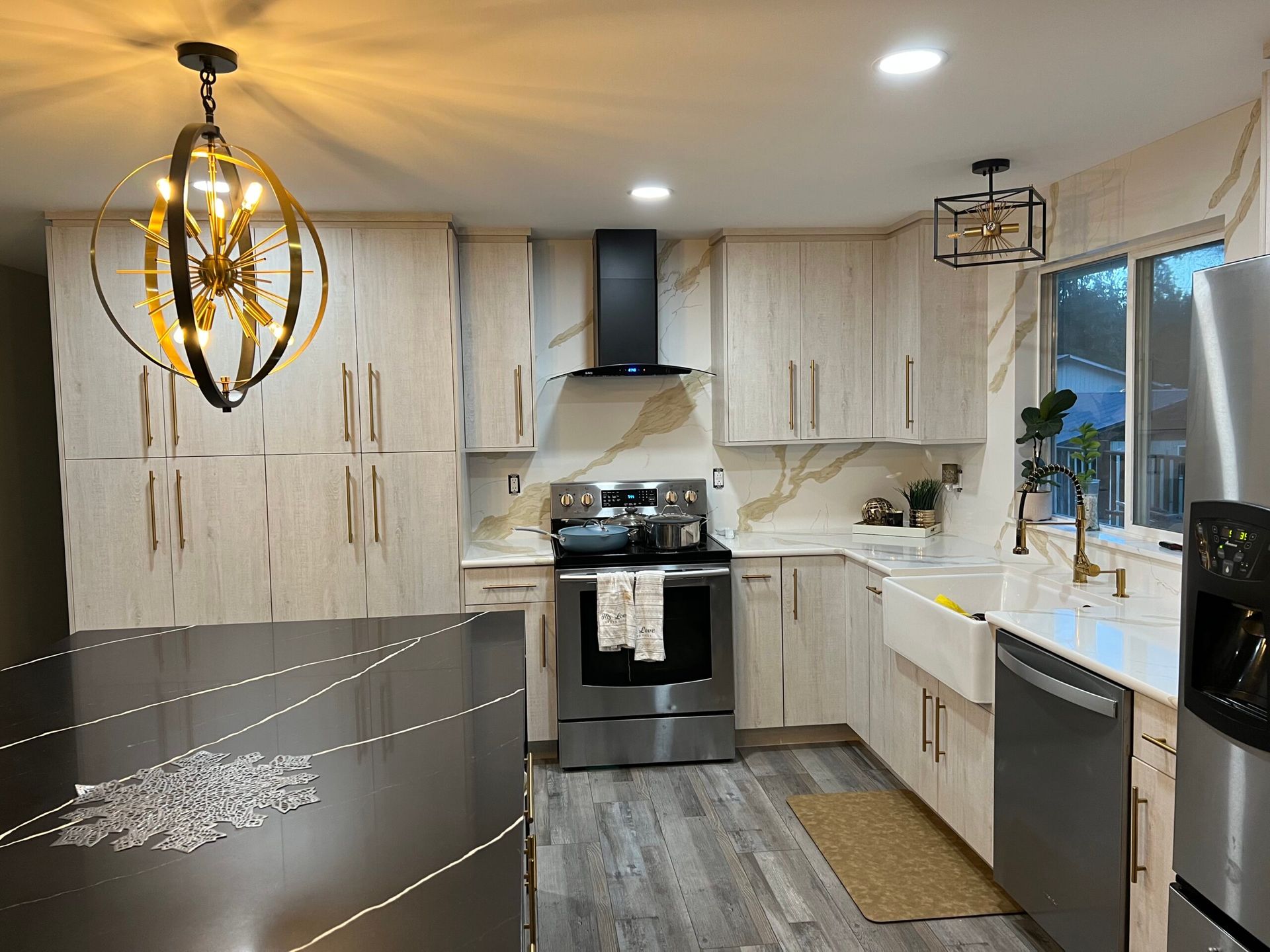 Modern kitchen with light wood cabinets, gold accents, stainless steel appliances, and a gray floor.