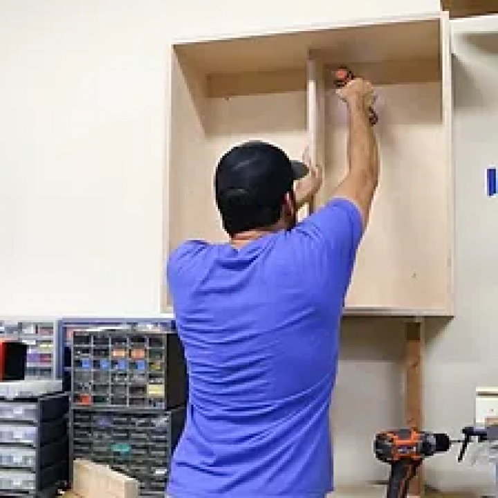 Person in blue shirt installing a cabinet shelf with a drill on a white wall.