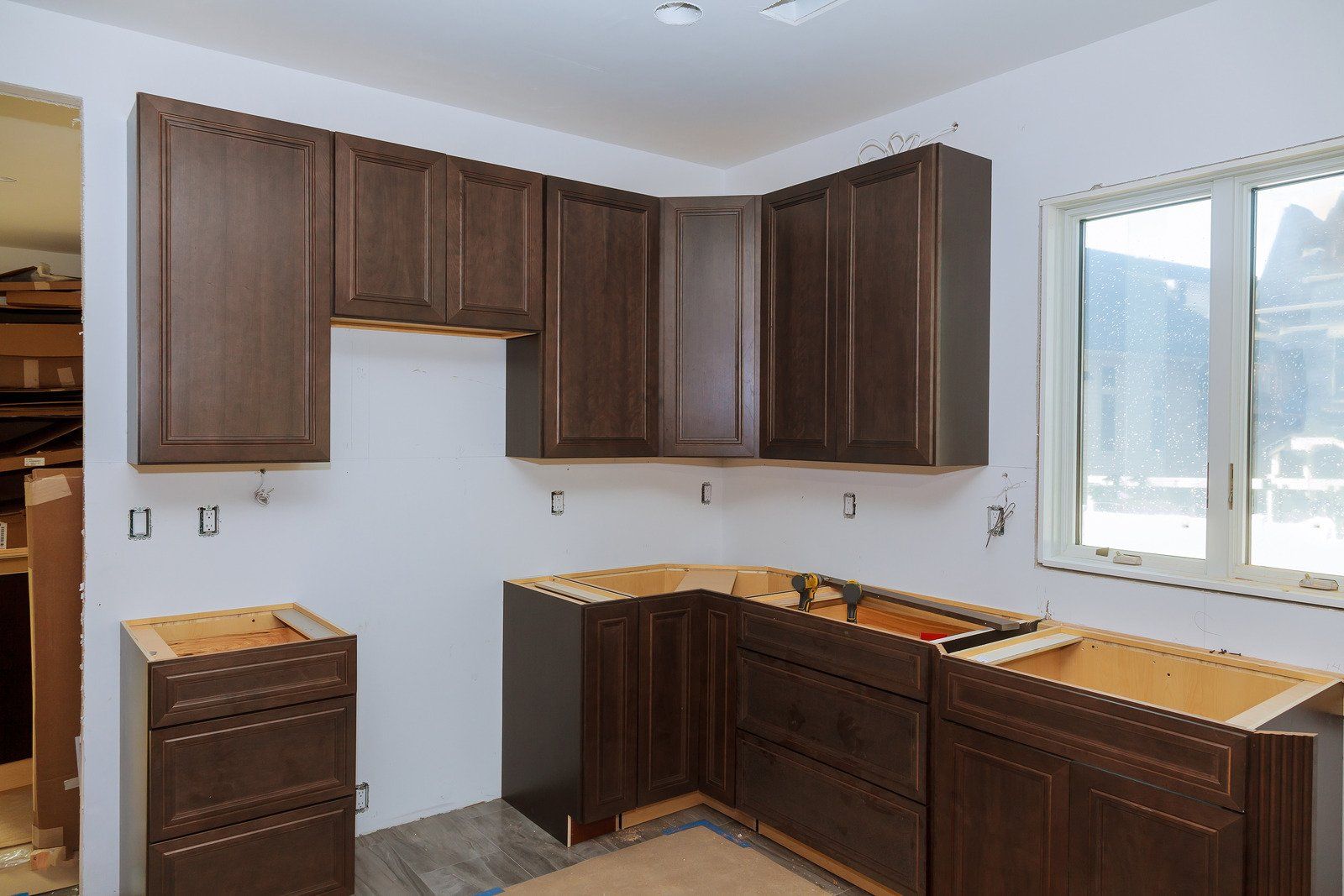 Kitchen under construction; brown cabinets, white walls, window, wood flooring.
