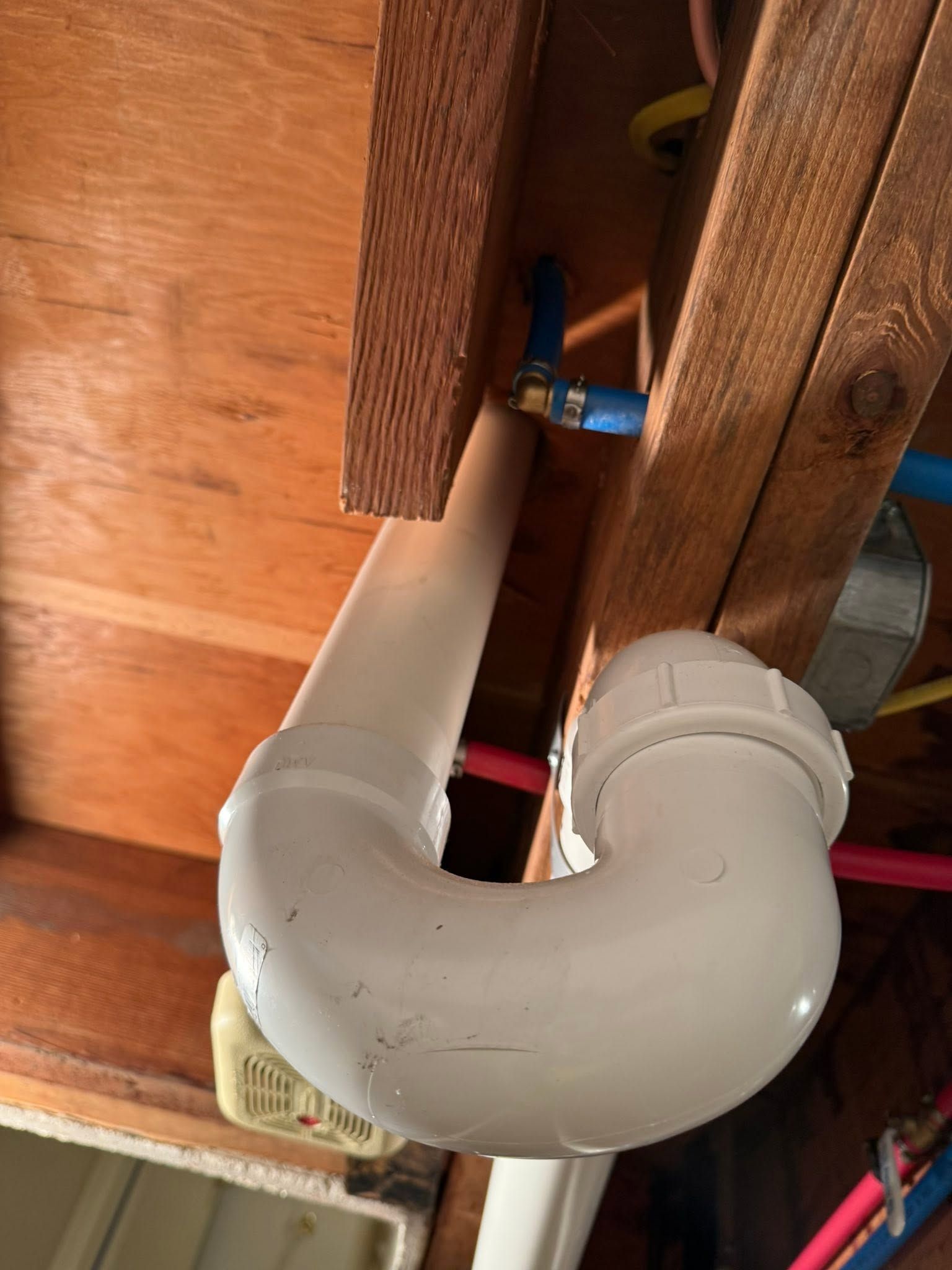 White PVC drain pipe elbow beneath wooden floor framing, with a blue valve and red strap.
