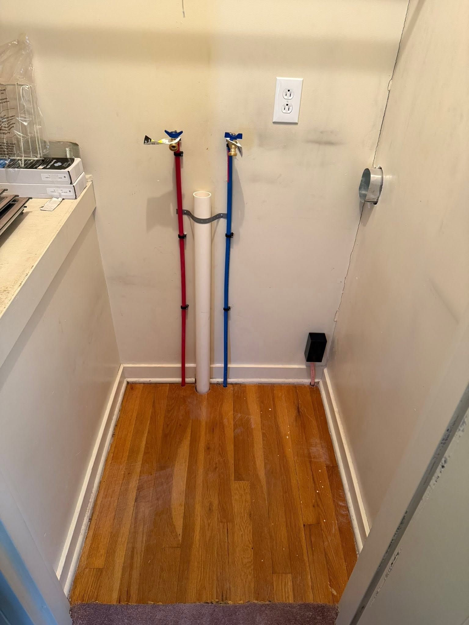Laundry hookup area with red and blue water lines, a drain, and wood-look flooring in a small utility space