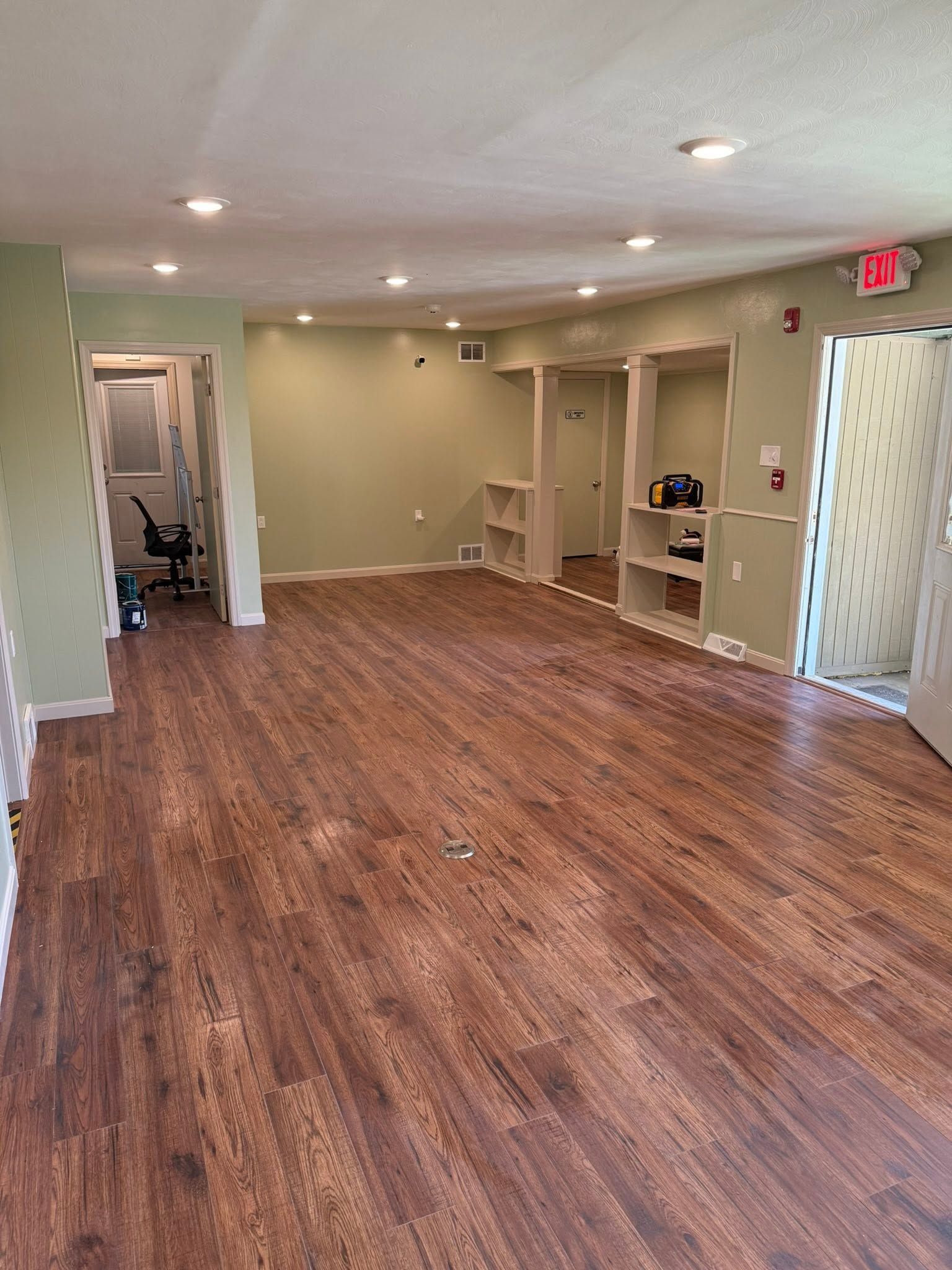 Empty room with wood flooring, recessed lights, built-in shelves, and a doorway with exit sign.
