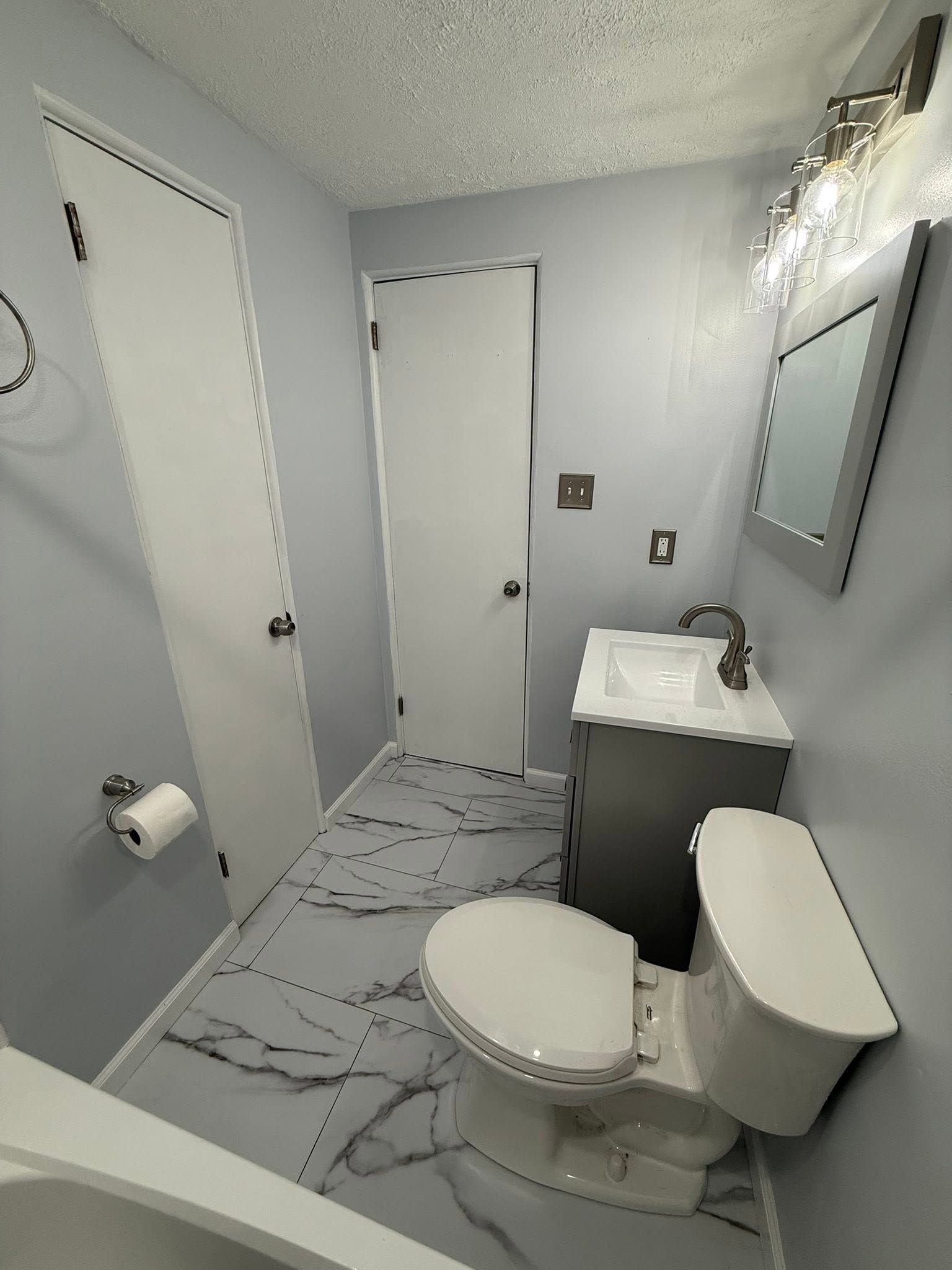 Small bathroom with white toilet, vanity sink, shower doors, and gray walls with marble-pattern floor.