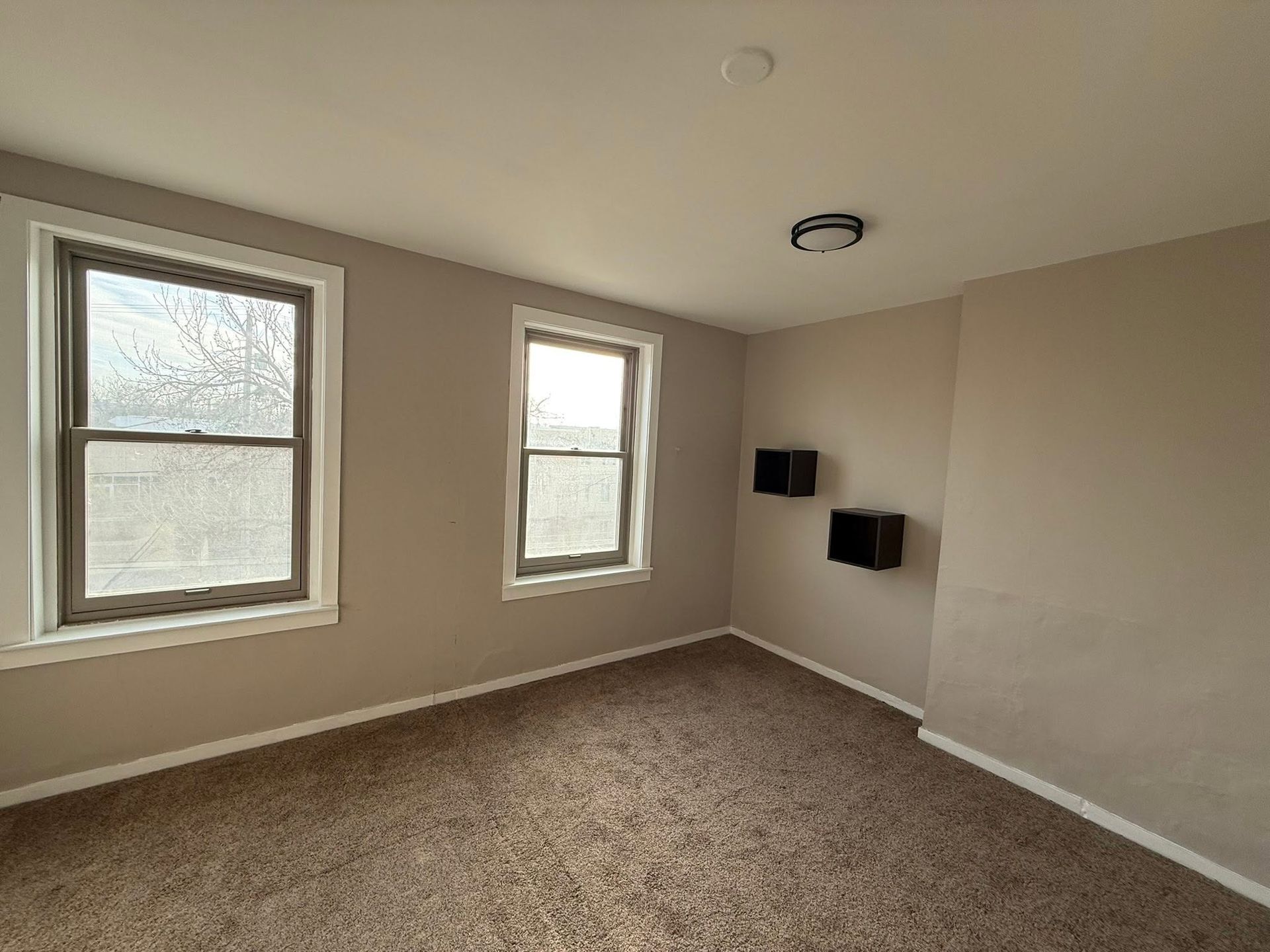 Empty beige bedroom with two windows, carpeted floor, and wall-mounted TV brackets.