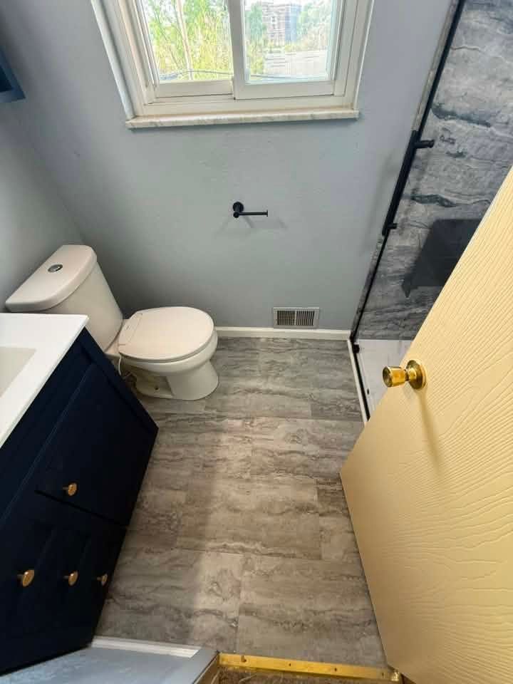 Small bathroom with toilet, vanity, window, and shower stall; gray walls and wood-look floor.
