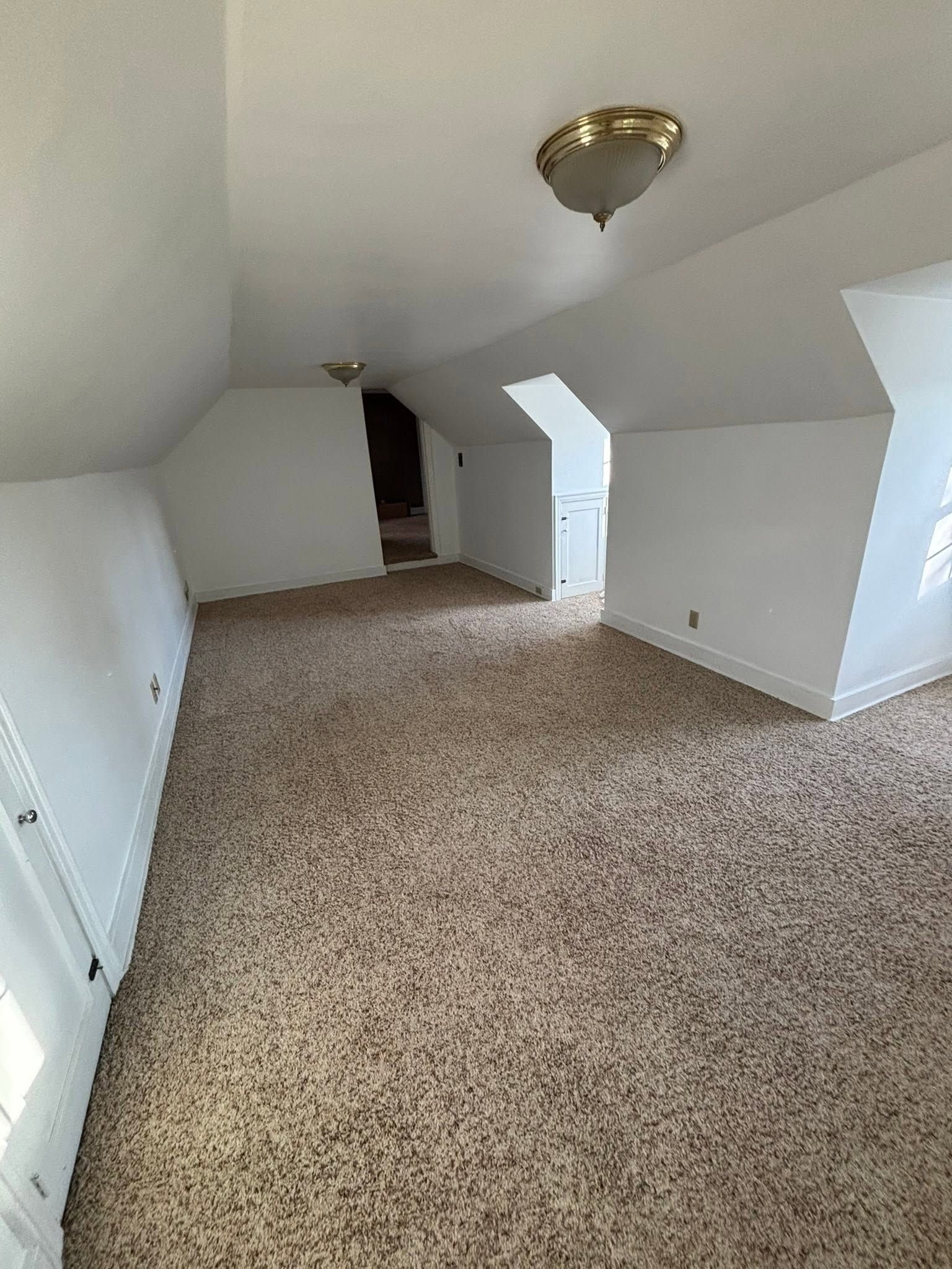 Empty carpeted attic room with white walls, sloped ceiling, and a brass ceiling light fixture.