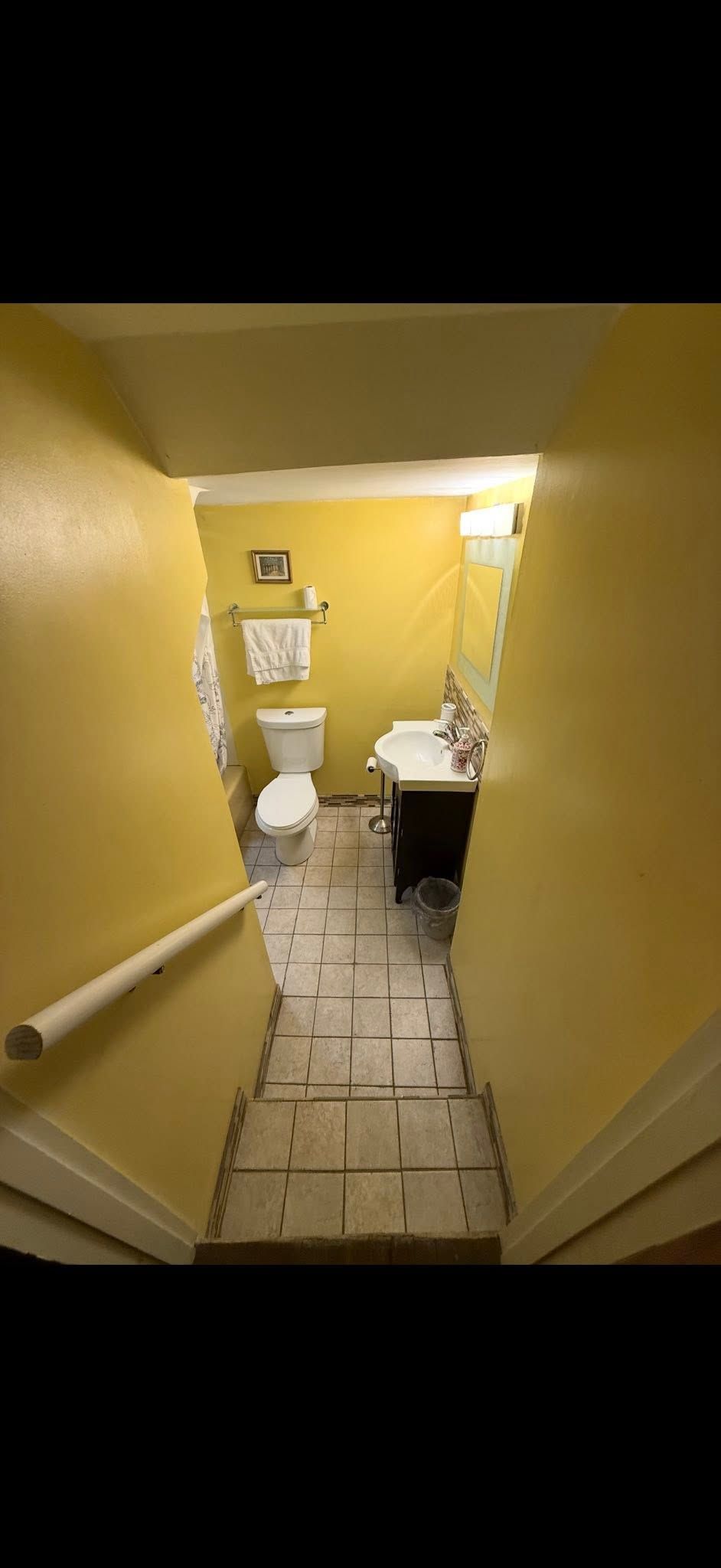Narrow yellow hallway leading to a small bathroom with a toilet, sink, and tiled floor.