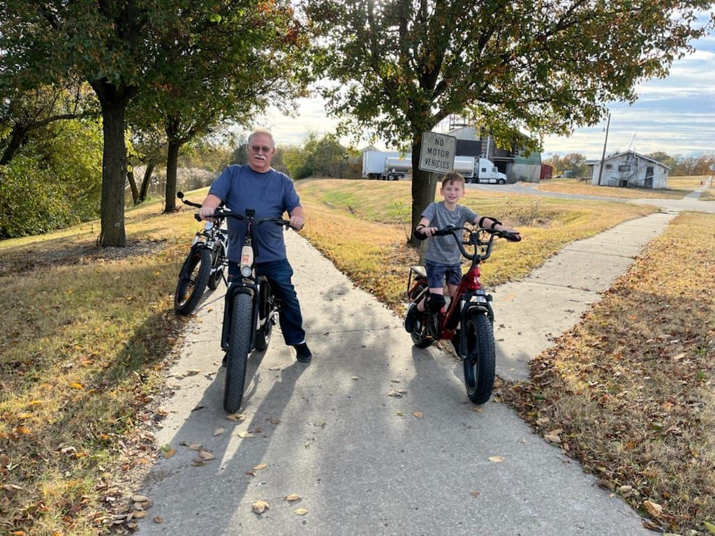 Man and child on electric bikes on a path; rural setting.