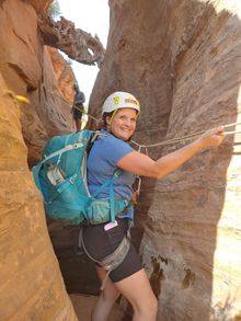 Woman in canyon, wearing helmet and harness, smiling while holding a rope.