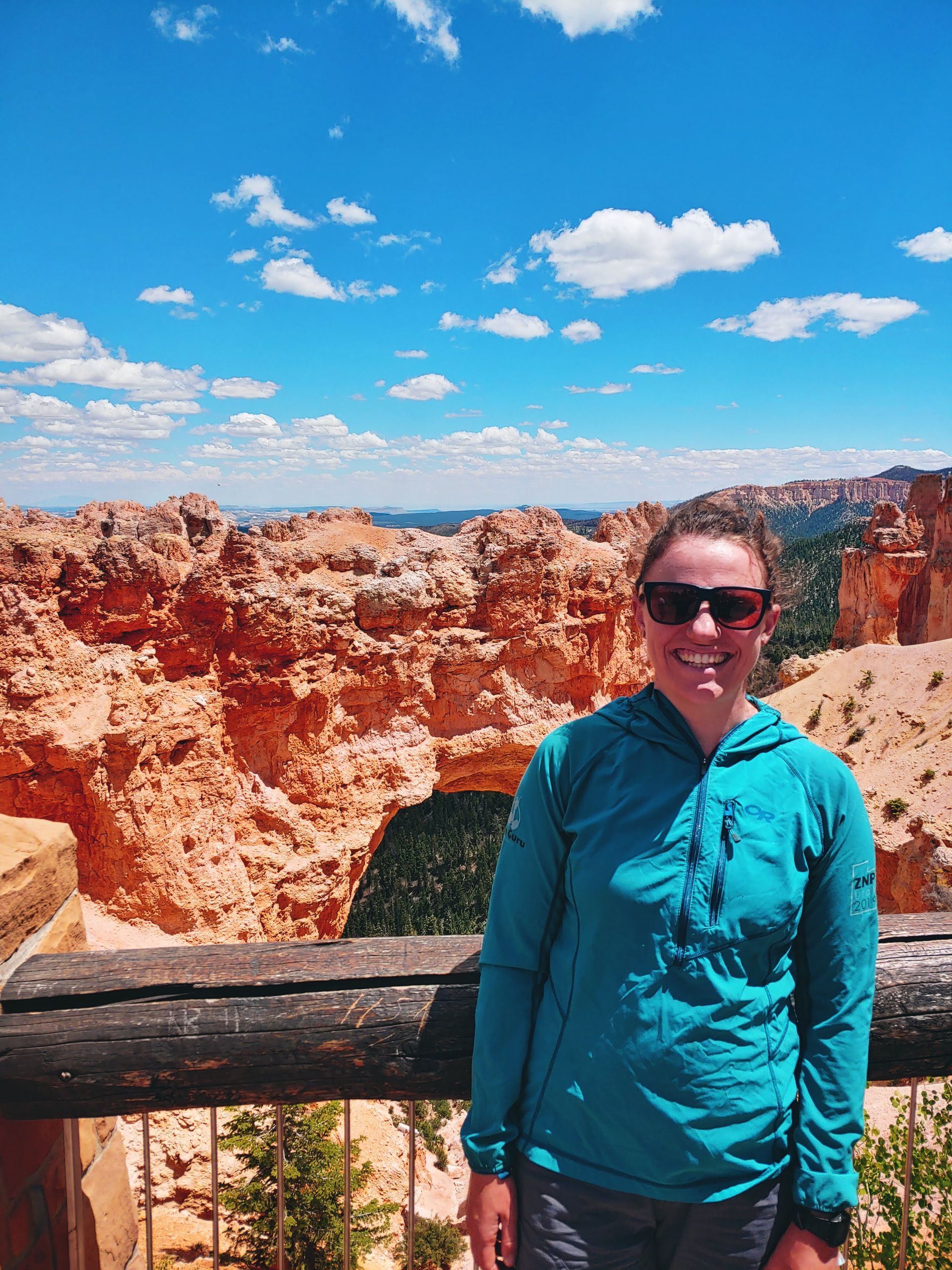 A woman in a blue jacket and sunglasses is standing in front of a canyon.